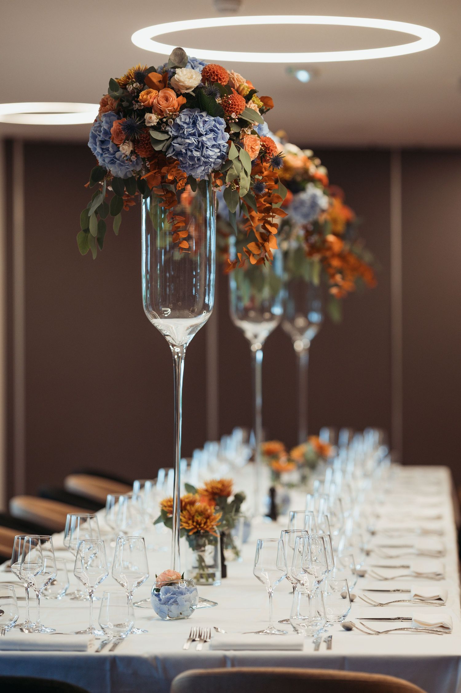 Elegant wedding reception table with tall glass vase centerpieces featuring blue hydrangeas and orange flowers.
