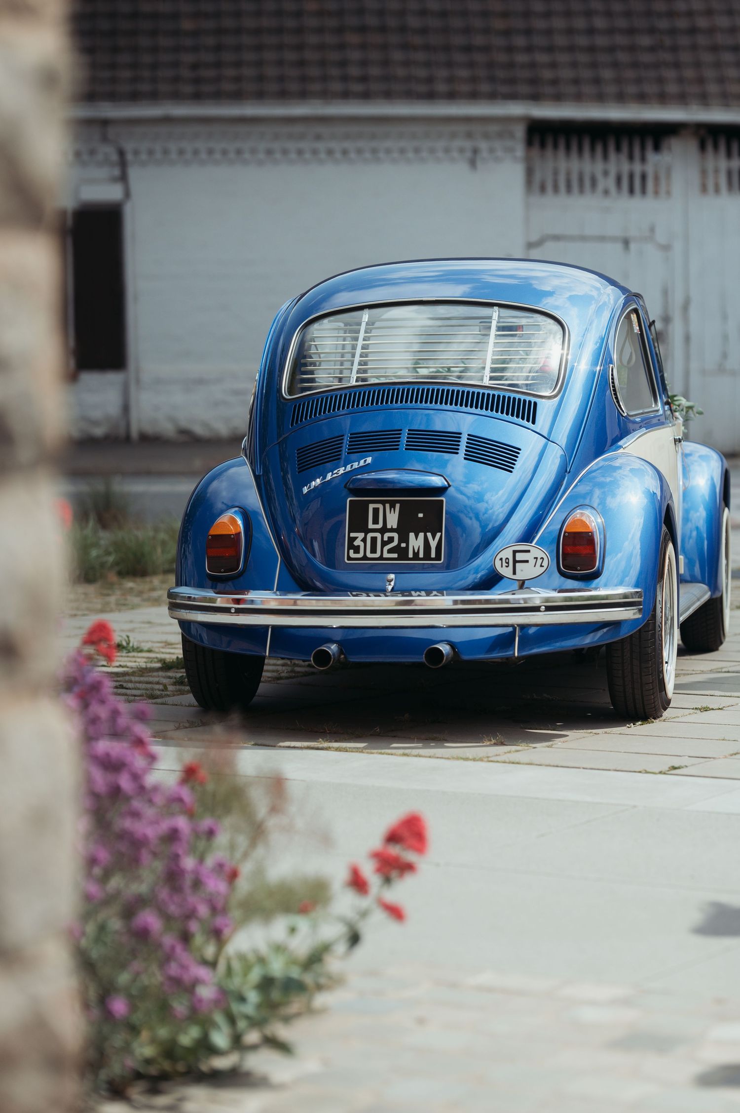 A classic blue Volkswagen Beetle with license plate DW 302-MY parked on a cobblestone street near colorful flowers.