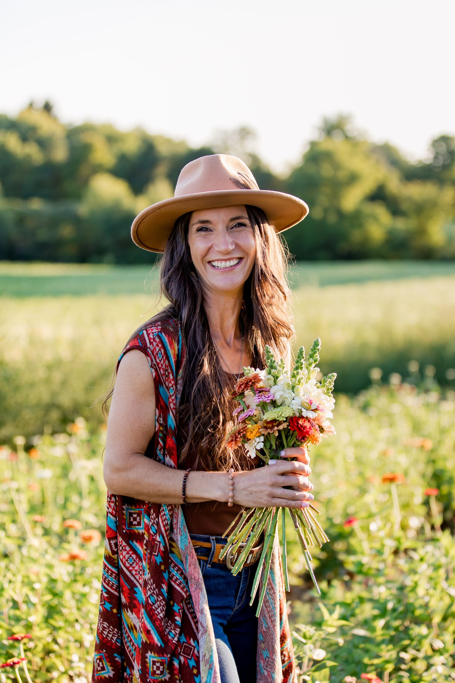 Female entrepreneur smiles in a cowboy hat and floral kimono holds freshly picked wildflowers in sunny summer field.