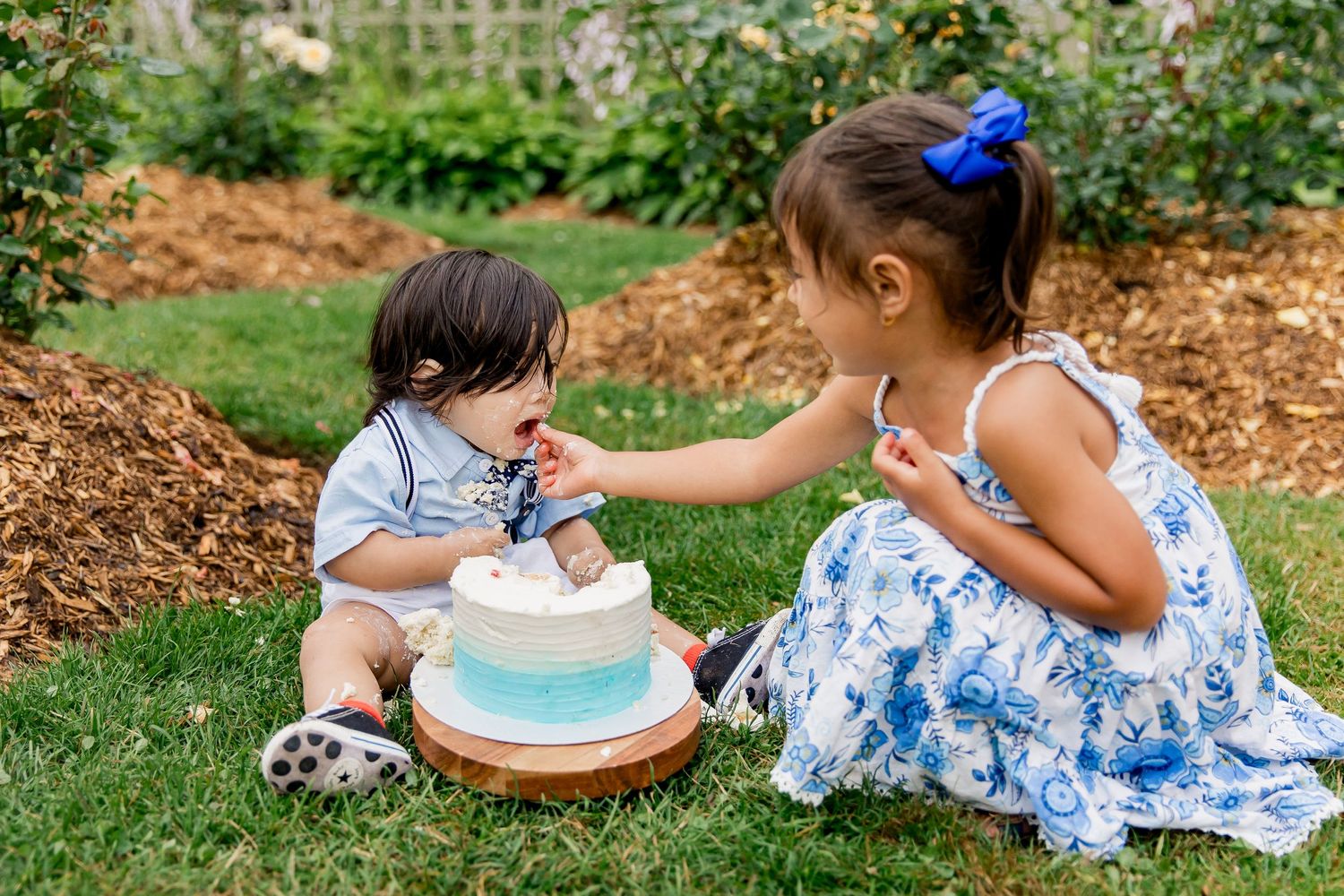 A brother and sister sitting on grass sharing a blue and white birthday cake at an outdoor cake smash photo session.