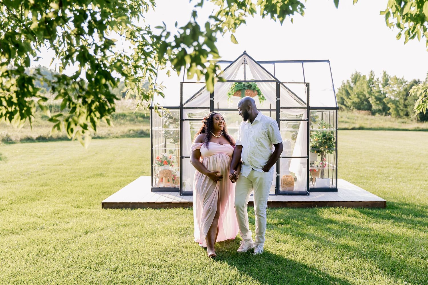 A couple walks in front of an elegant glass greenhouse structure on a green lawn during a maternity session.