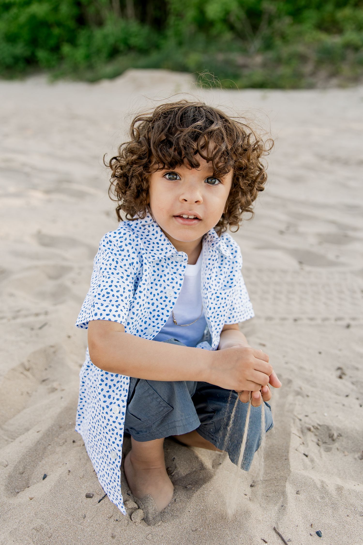 Child in blue polka dot shirt and denim shorts crouching on sandy beach with curly hair and natural lighting playing with the sand.