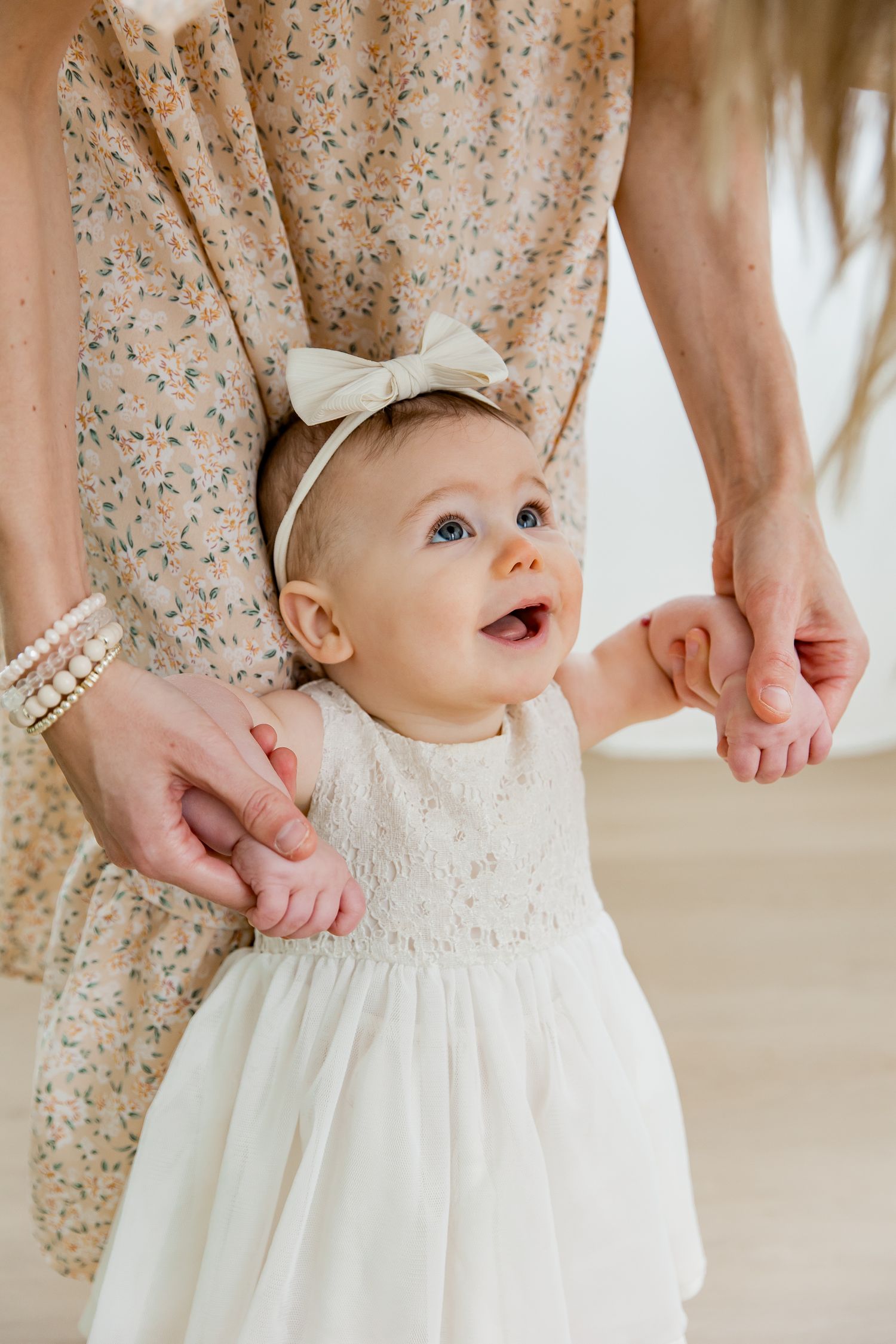 A baby wearing a white lace dress and bow headband smiles while being held up by mommy's hands in a softly lit room.