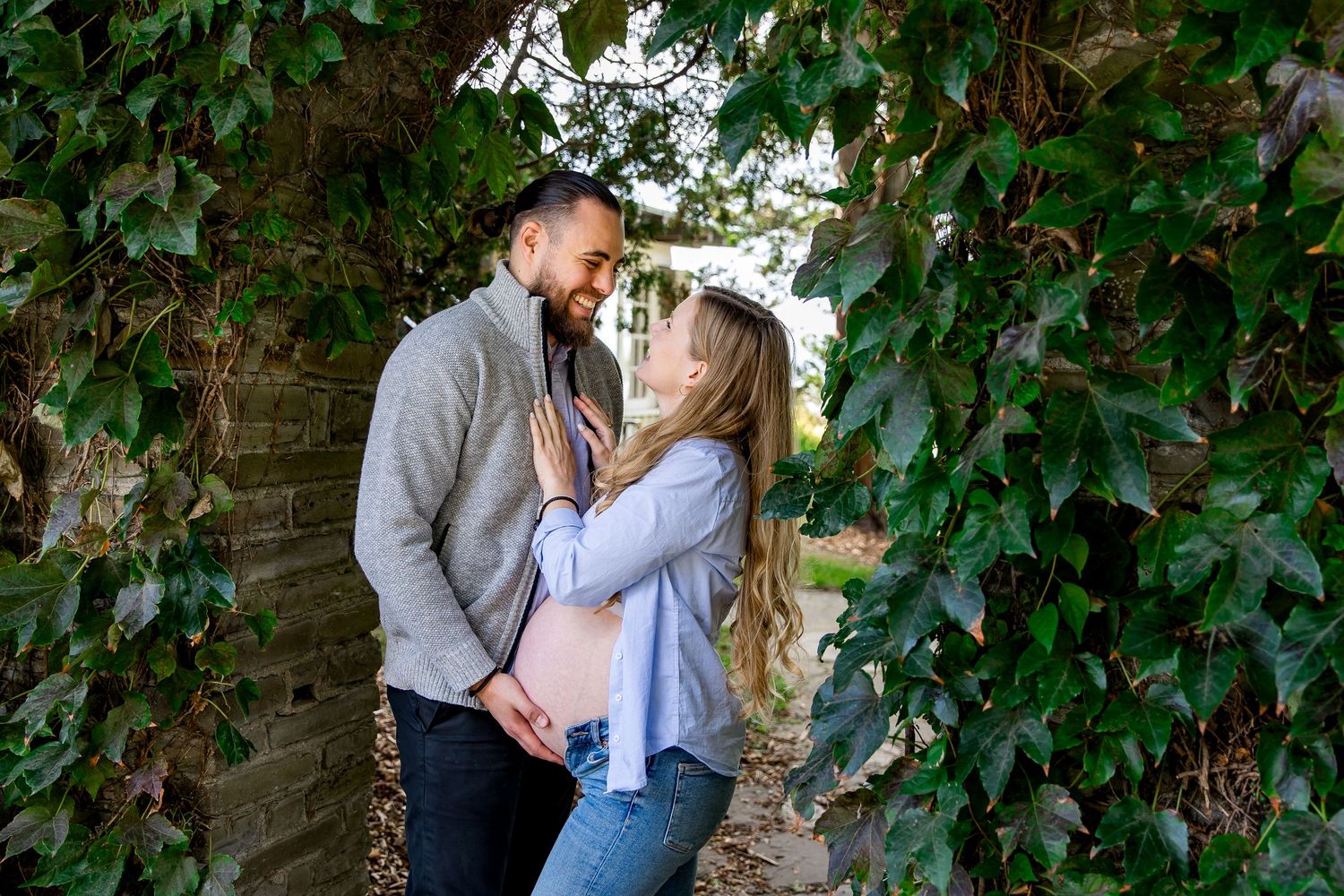 Romantic couple embracing against ivy-covered wall during outdoor maternity photoshoot.