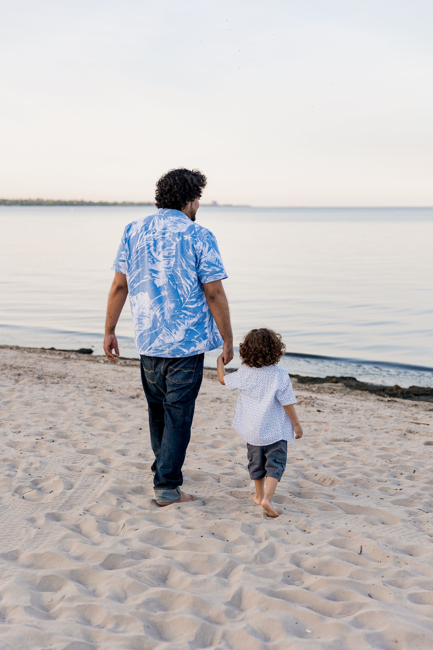An father and child walking together on a sandy beach at sunset wearing casual summer clothing.