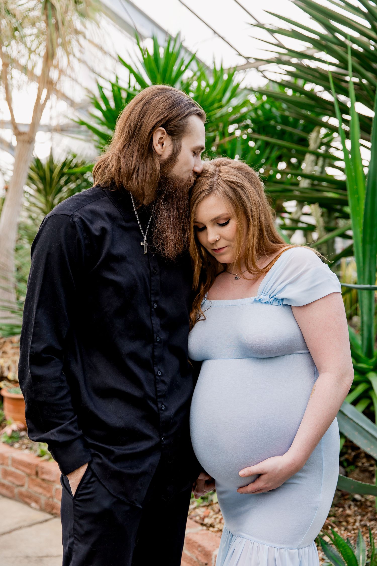 A couple share an intimate moment in front of tropical plants, with her wearing a light blue maternity dress.