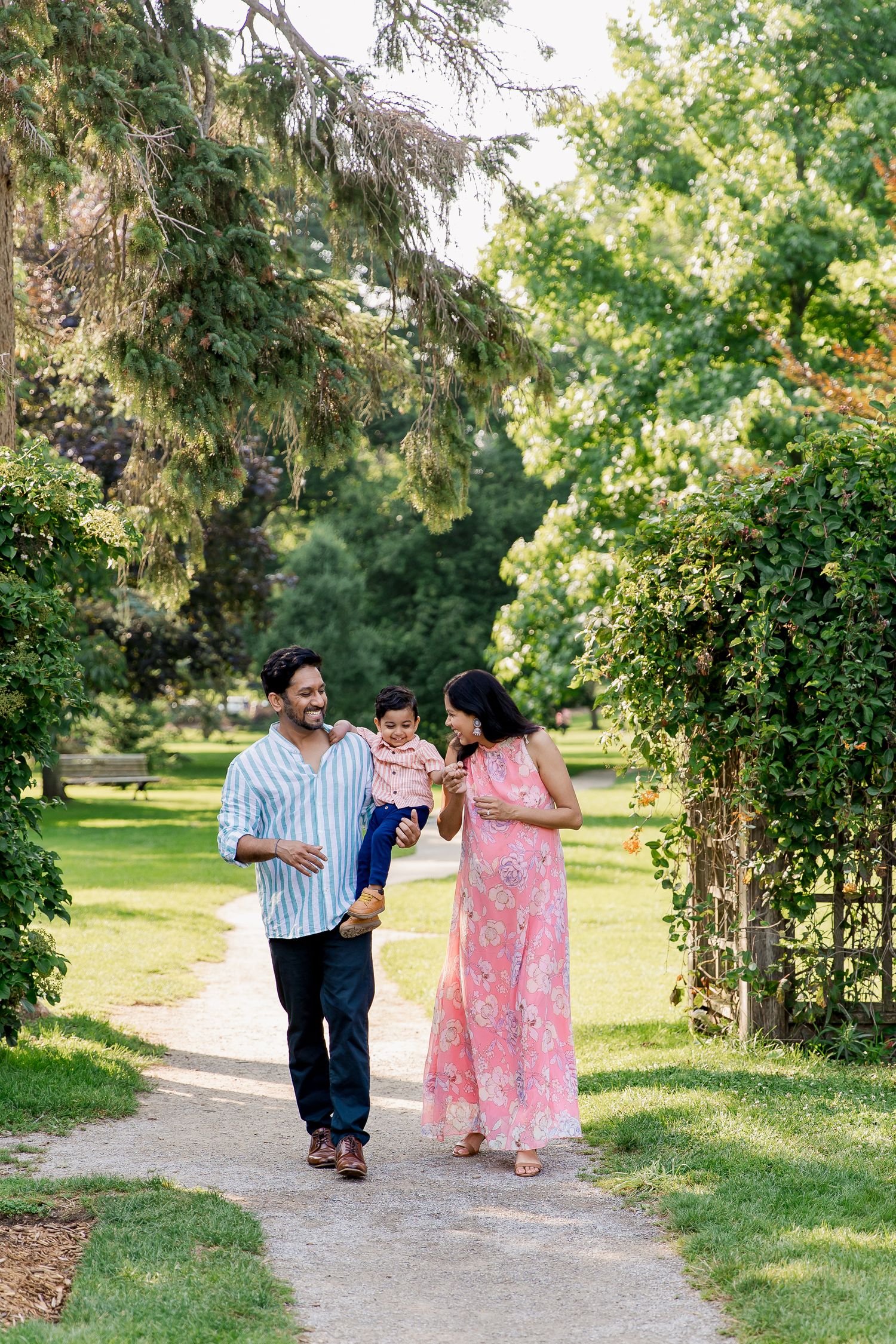 A family walks together on a garden path under lush green trees and flowering archways on a sunny summer day.