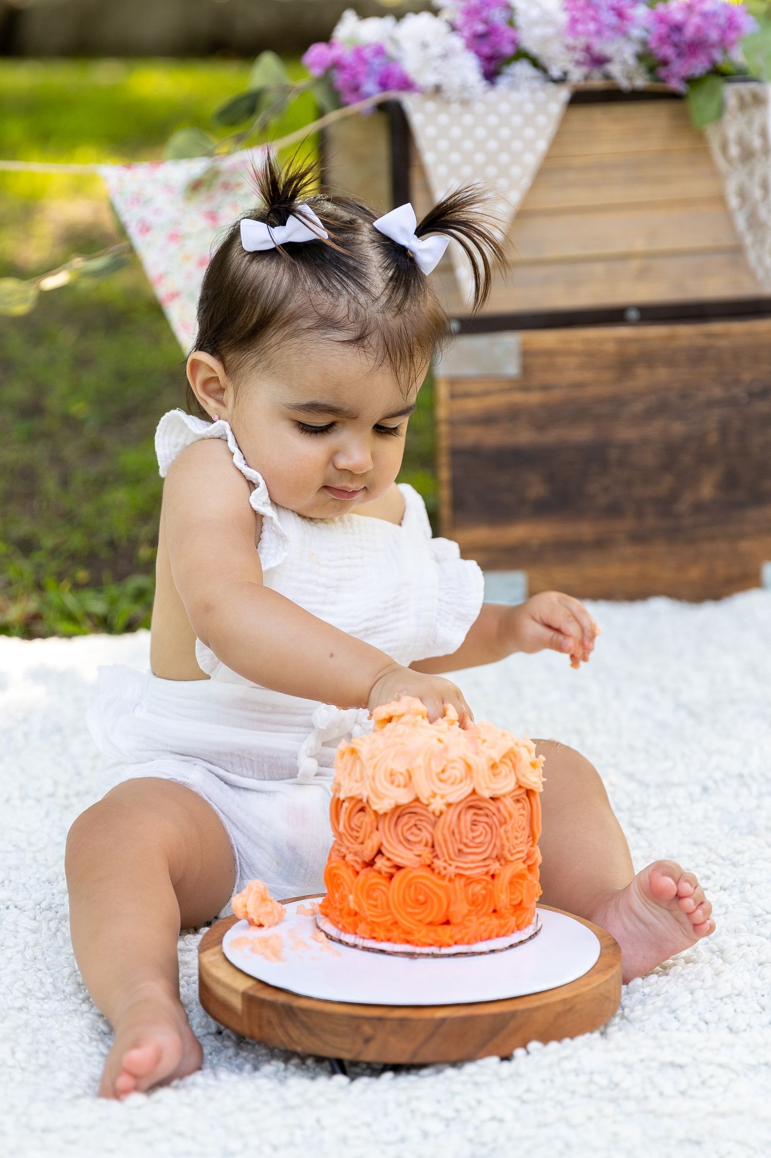 A small child in a white outfit sits on a blanket reaching for an orange ombre cake decorated with roses for her outdoor cake smash photos.