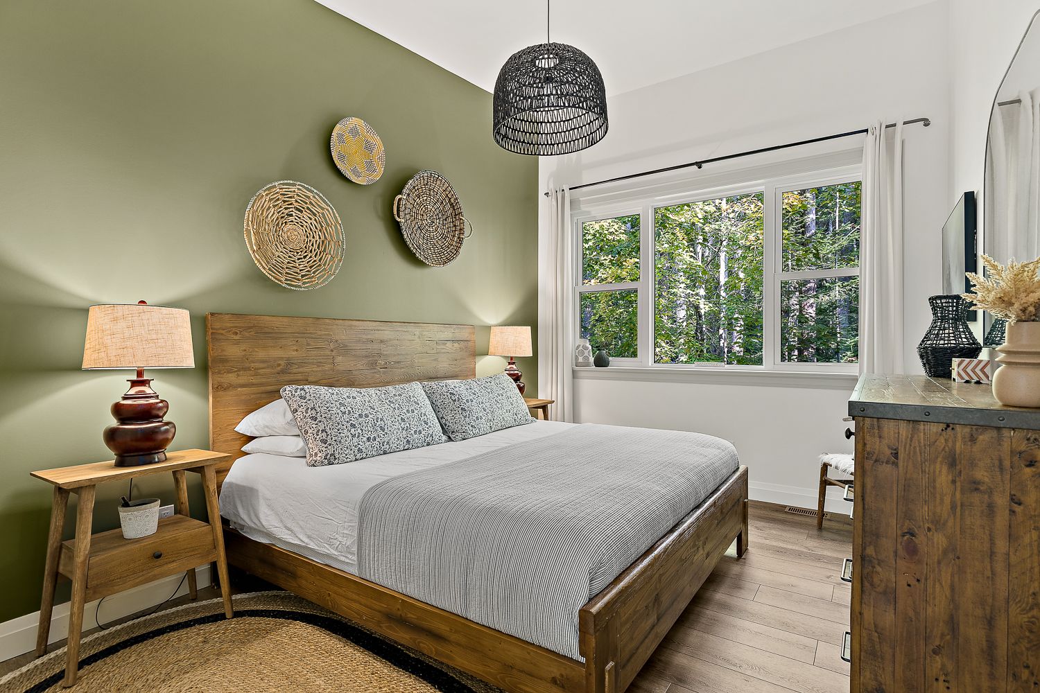 Cozy Muskoka AirBnB bedroom with wood bed frame, green feature wall, decorative wall art, and large window overlooking trees.