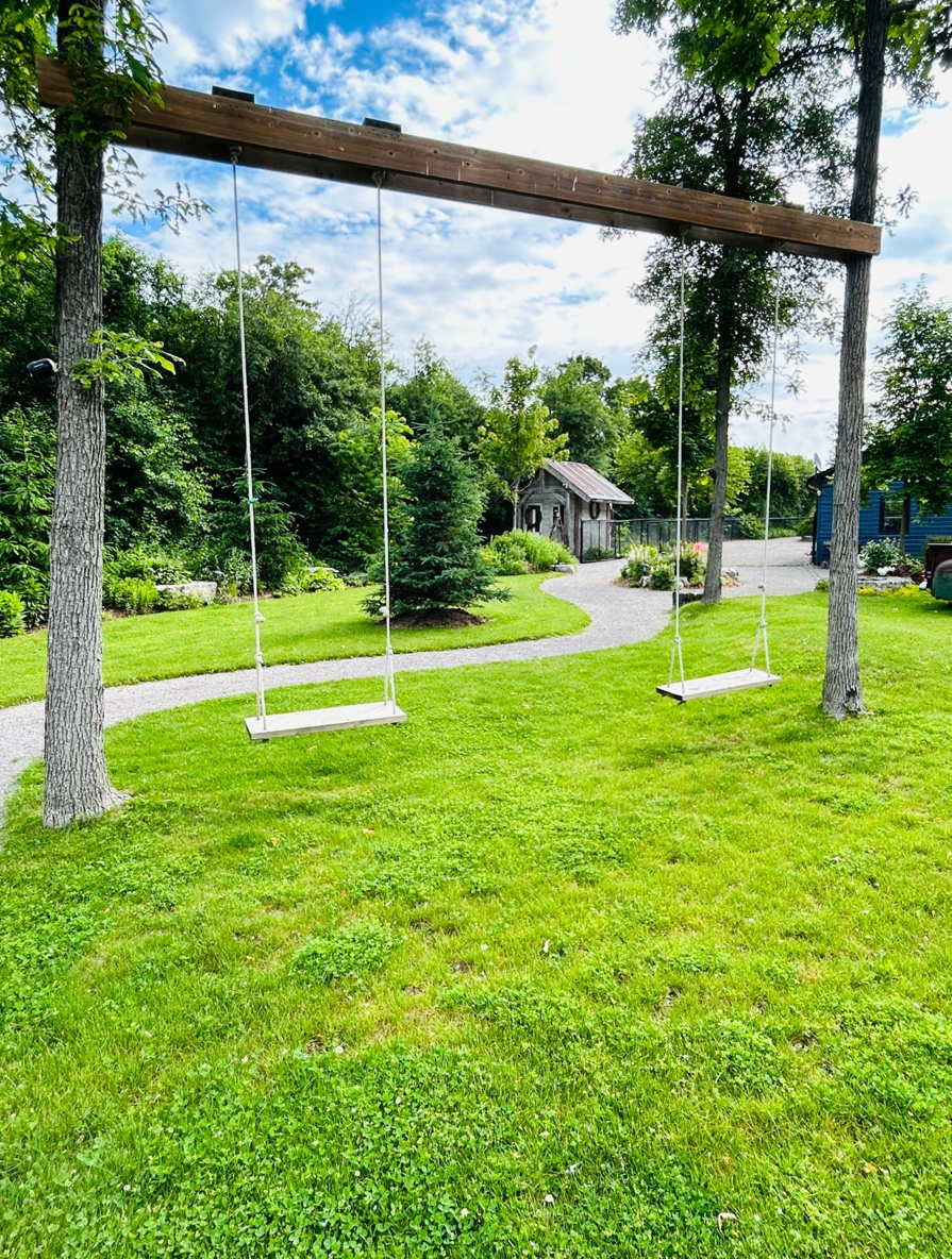A rustic wooden swing set with two swings sits on a green lawn with a winding path and cabin bunkie in the background.