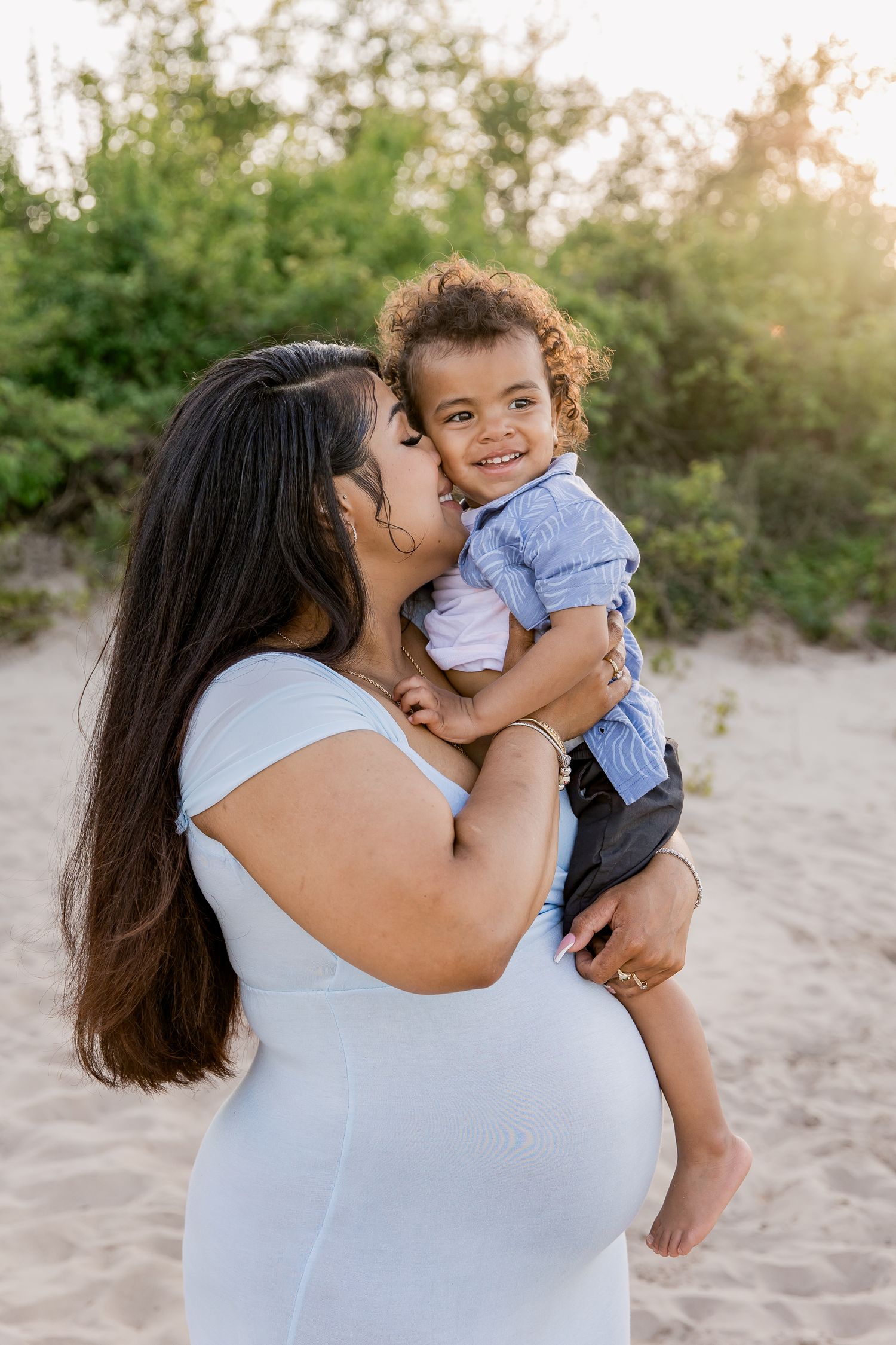 A expectant mother in a light blue dress holds a small child wearing blue against a sunset beach backdrop.