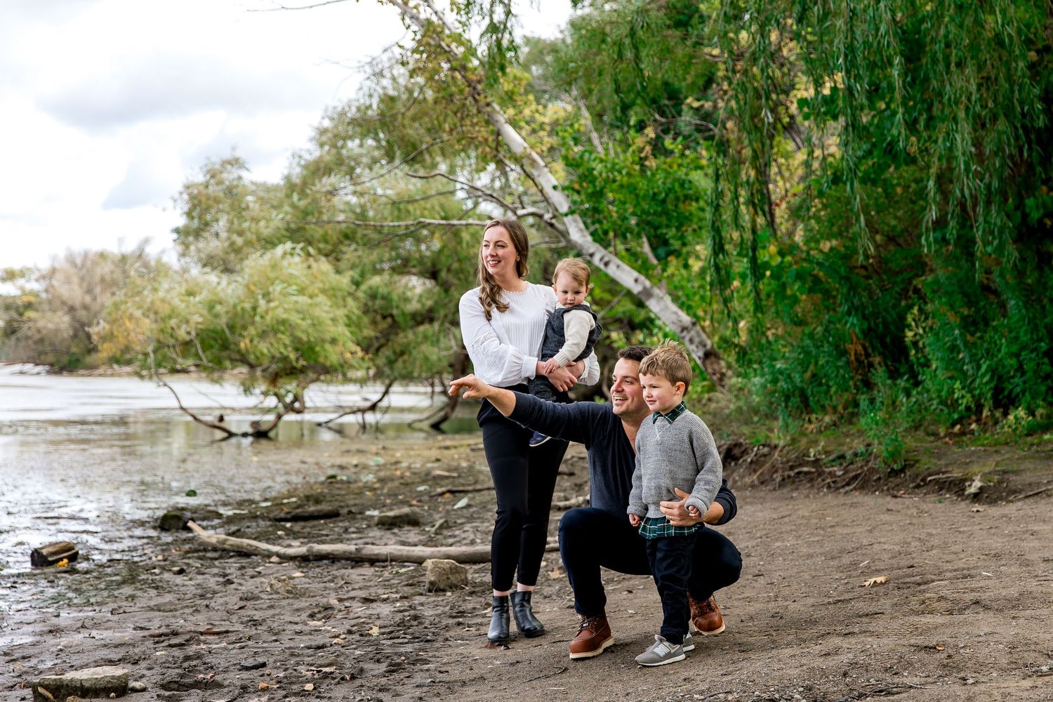 Candid family photo session happily skipping rocks together on a scenic riverbank with willow trees and water in the background.