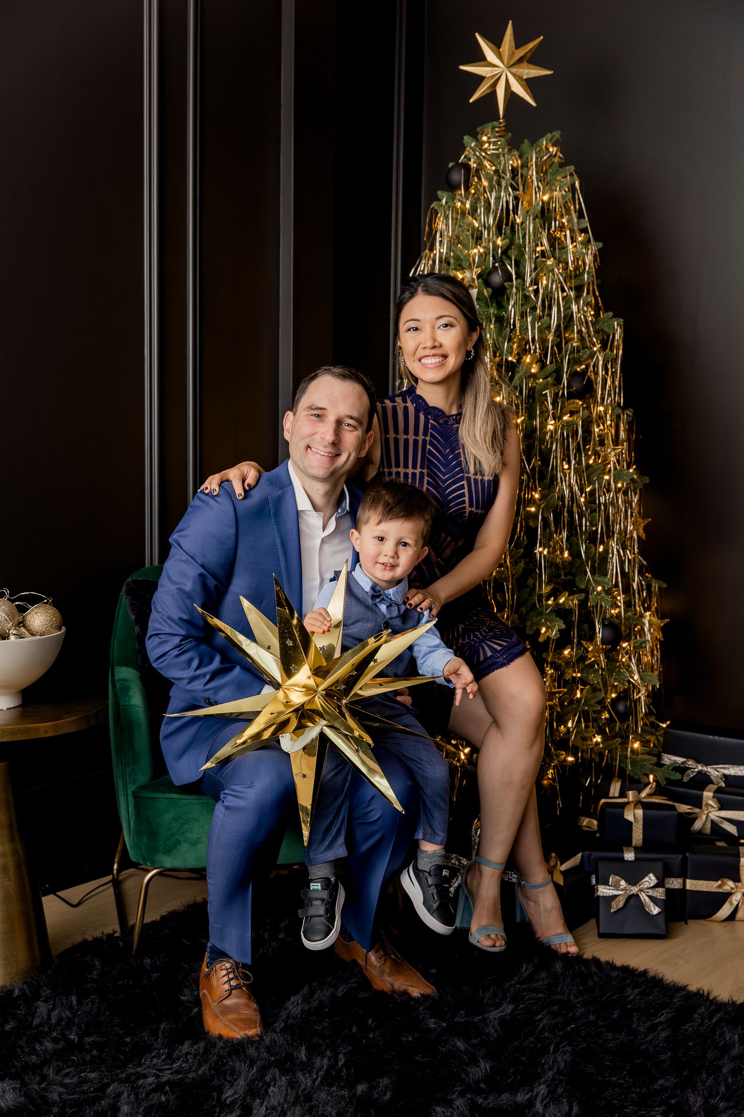 Family poses together near an elegantly decorated gold Christmas tree with a star on top in a dark room with plush carpet.