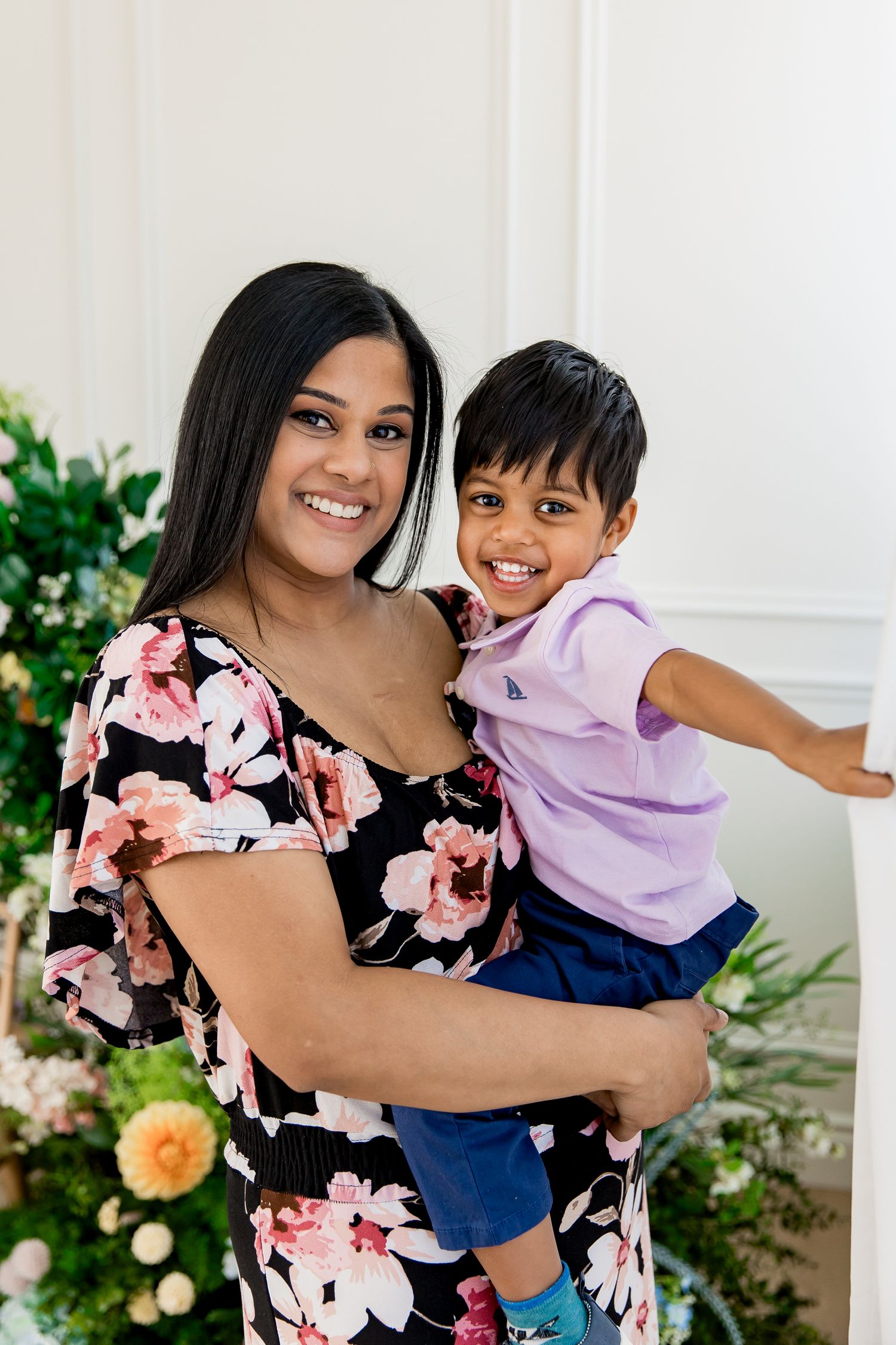 Mom and toddler son share a joyful moment in front of a floral installation, with one wearing a floral dress and one in lavender golf shirt.