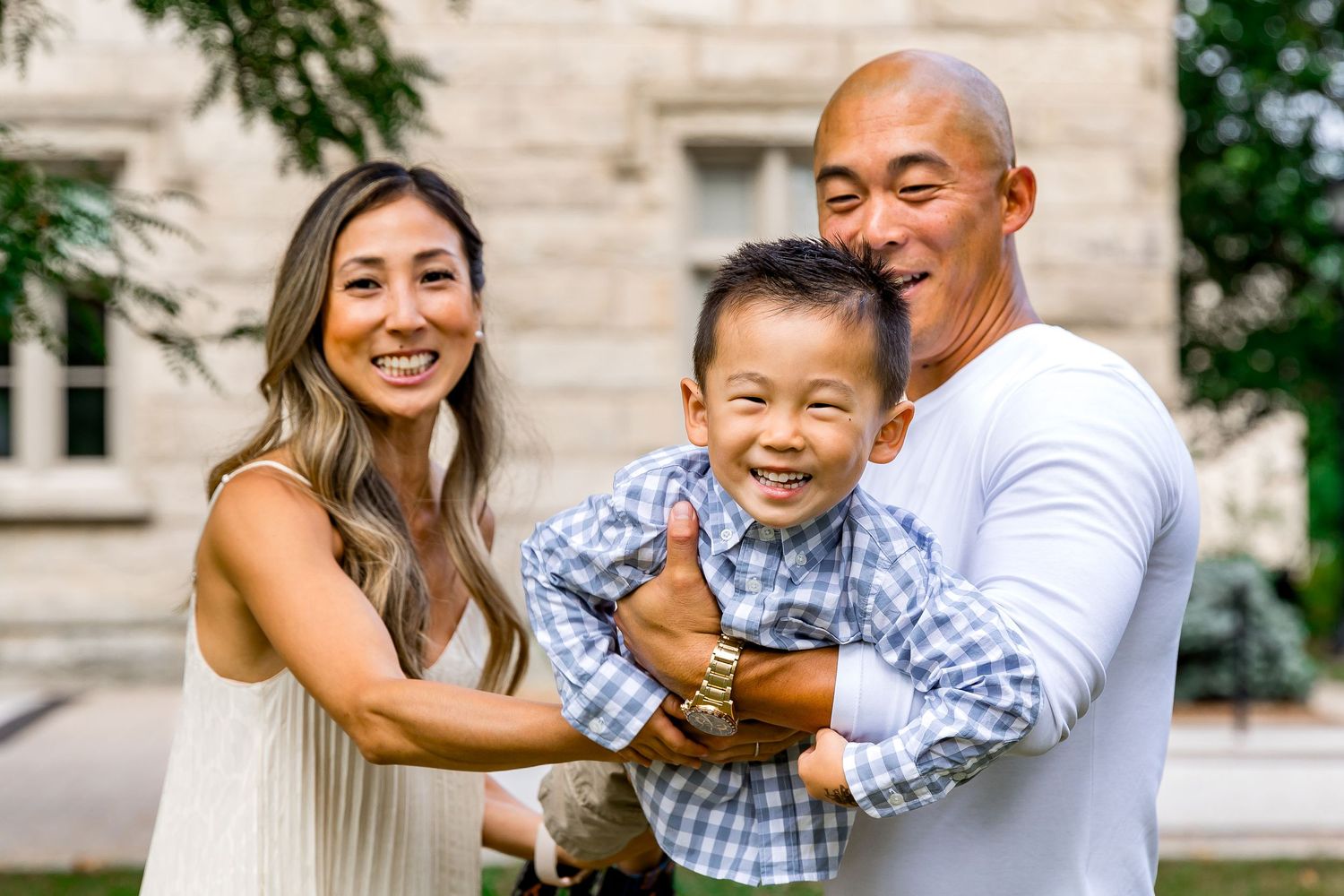 Parents of a toddler swing him in their arms having fun in front of a historical building in downtown Milton for their family photo session.