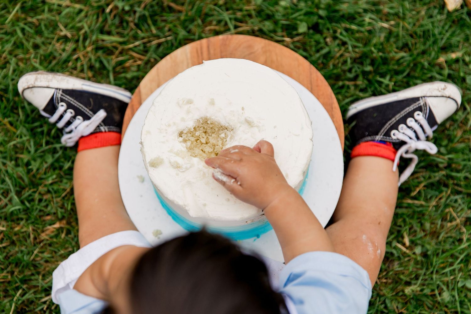 An overhead view of a child sitting on grass eating cake wearing black Converse style sneakers for his outdoor cake smash photo session.