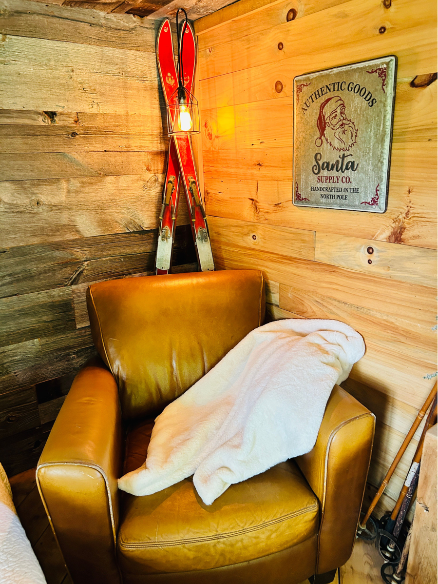 Vintage red skis lean against wooden cabin wall near brown leather chair with white blanket and retro Santa sign.