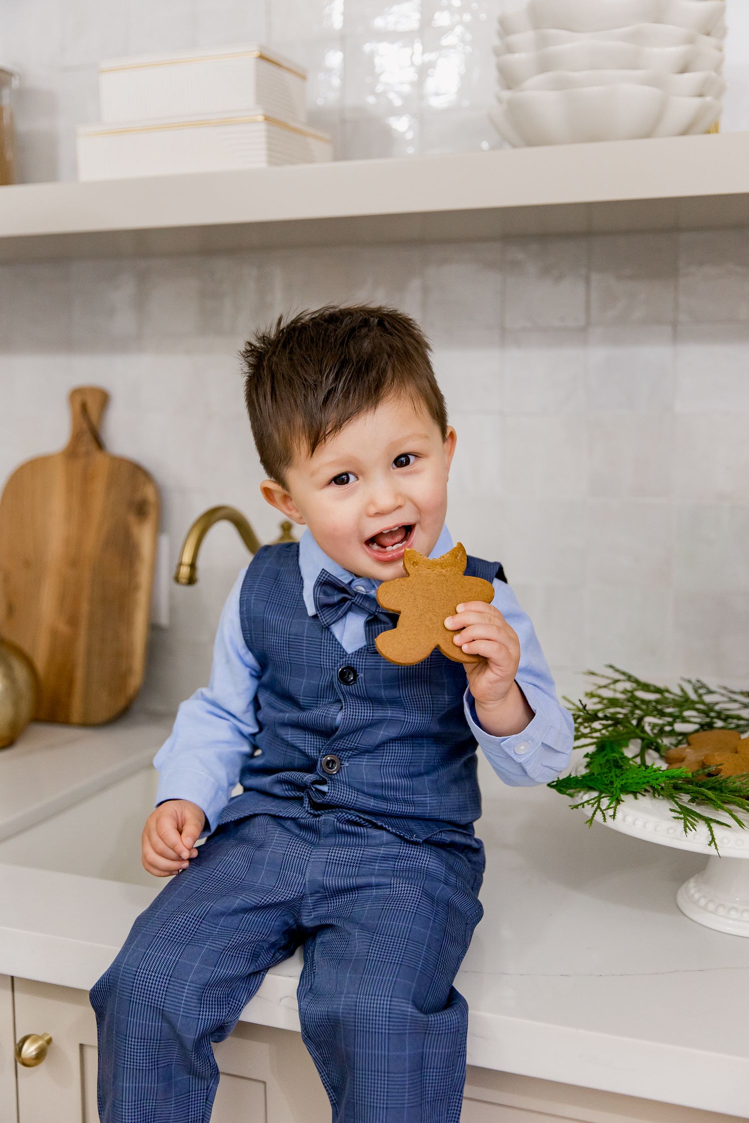 A happy child sits on a kitchen counter wearing a navy suit and light blue shirt while enjoying a gingerbread cookie.