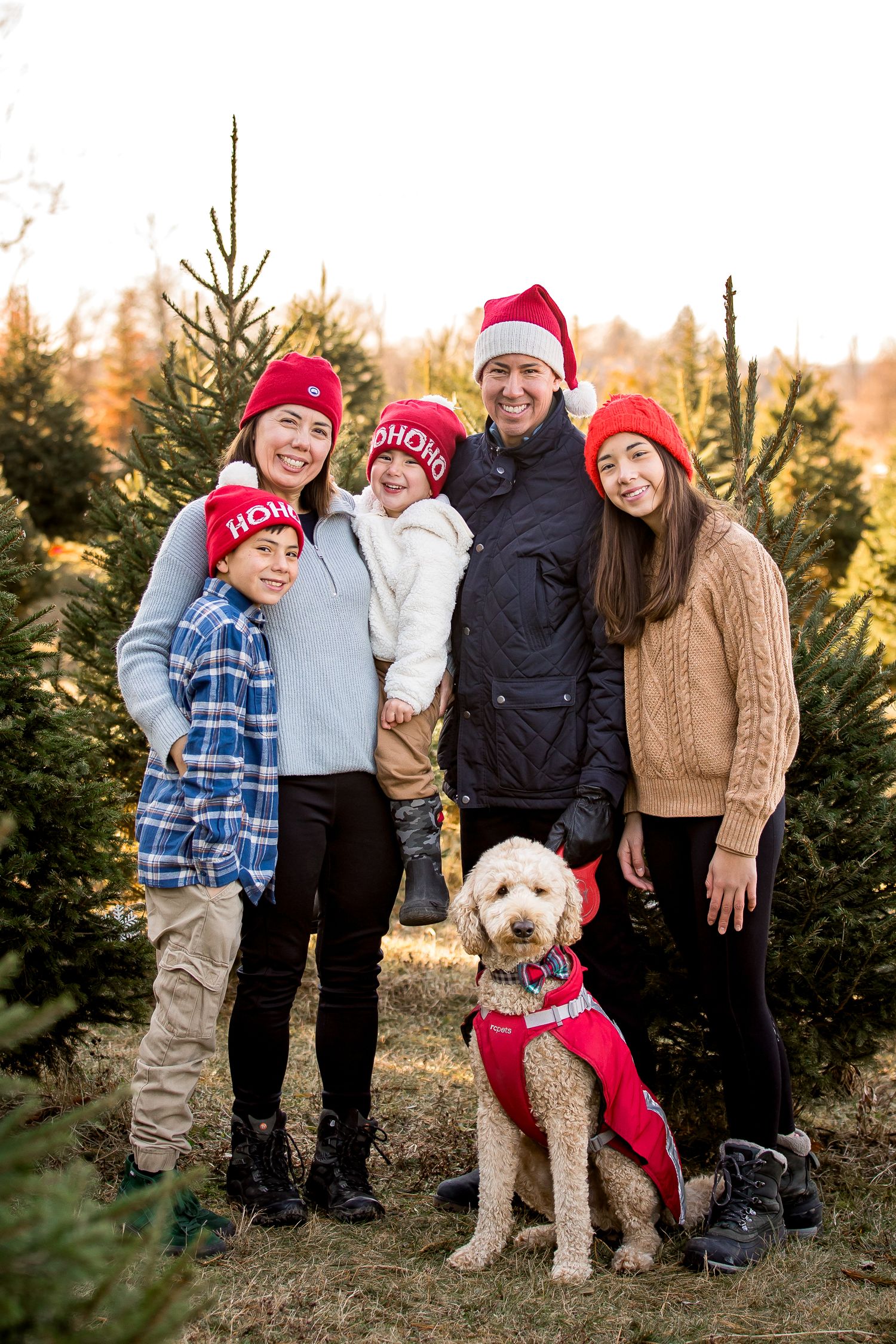 A festive family wearing red winter hats stands among evergreen trees with their goldendoodle dog during Christmas season.