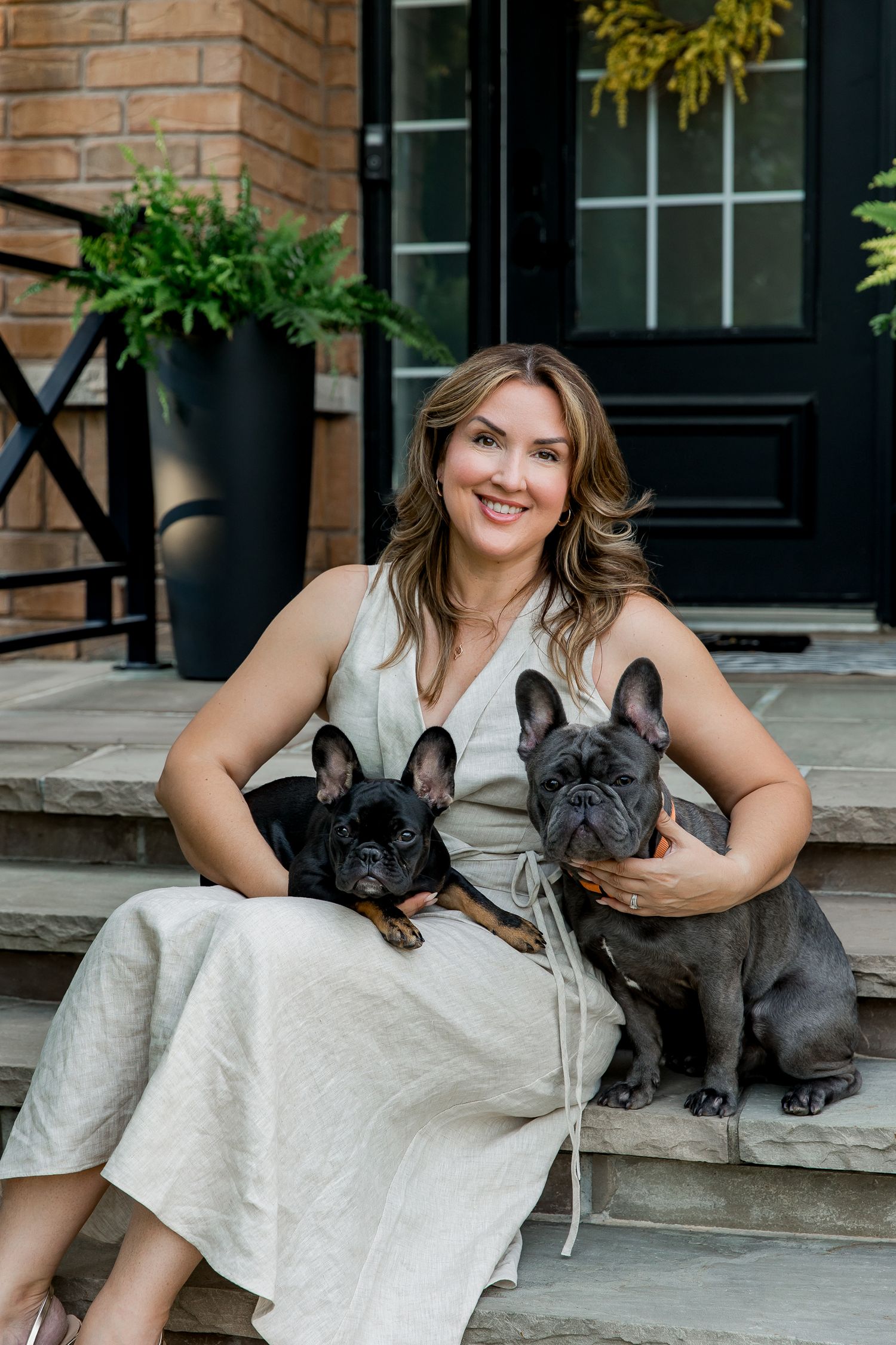 Two Frenchies sit with someone in a white dress on front porch steps with potted plants and black door.