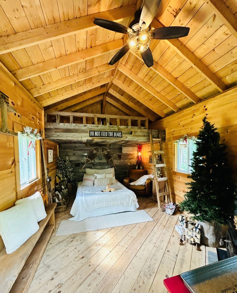 Cozy rustic cabin interior with wooden walls, Christmas tree, white bed, and ceiling fan in vaulted pine ceiling.
