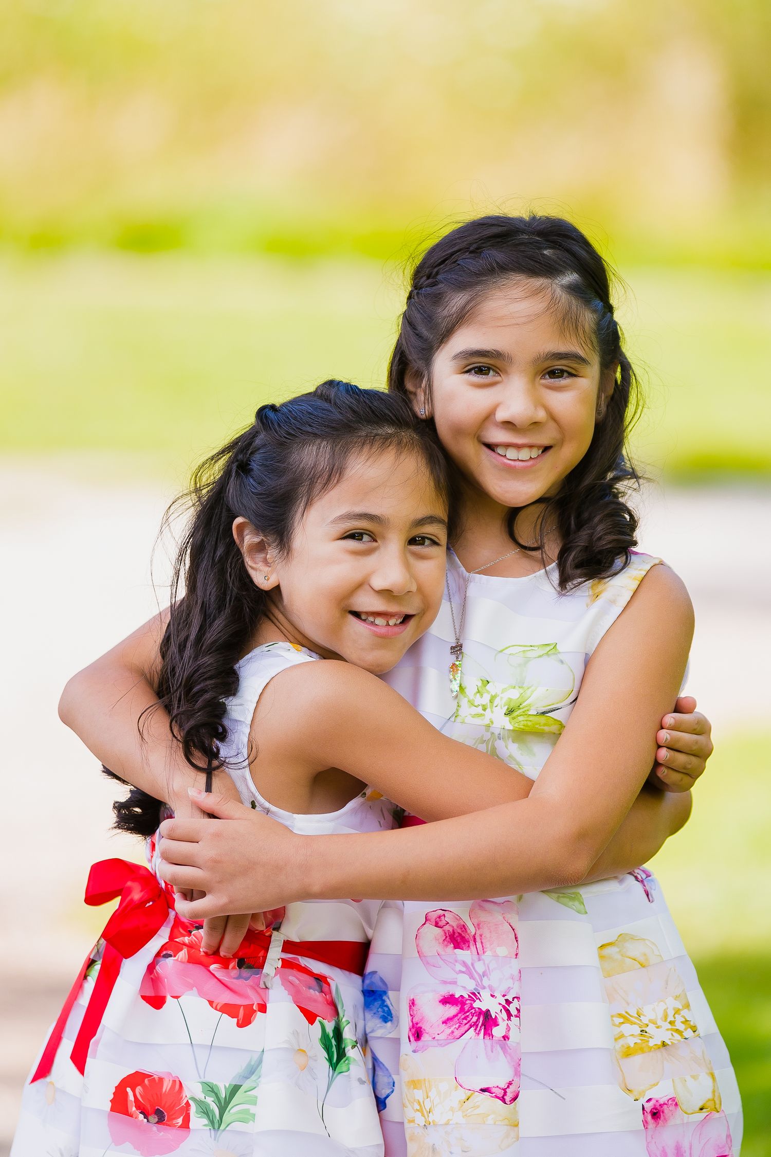 Two young sisters in floral summer dresses share a happy embrace outdoors on a sunny day.
