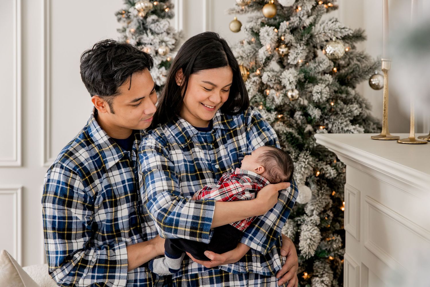 Cozy family scene with matching plaid pjs next to decorated Christmas tree and neutral fireplace mantel.