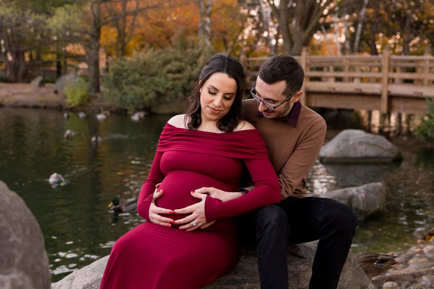 A couple sits by a pond in autumn, with the pregnant woman wearing a burgundy dress while her partner embraces her lovingly.