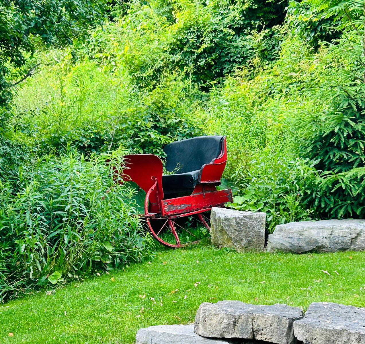 A vintage red sleigh chair sits among lush greenery and stone steps in a garden setting.