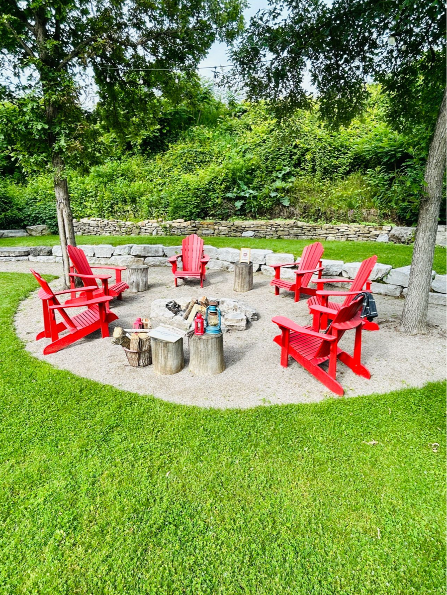 Red Adirondack chairs arranged in a circle around a fire pit on paved stone patio area surrounded by lush green lawn.