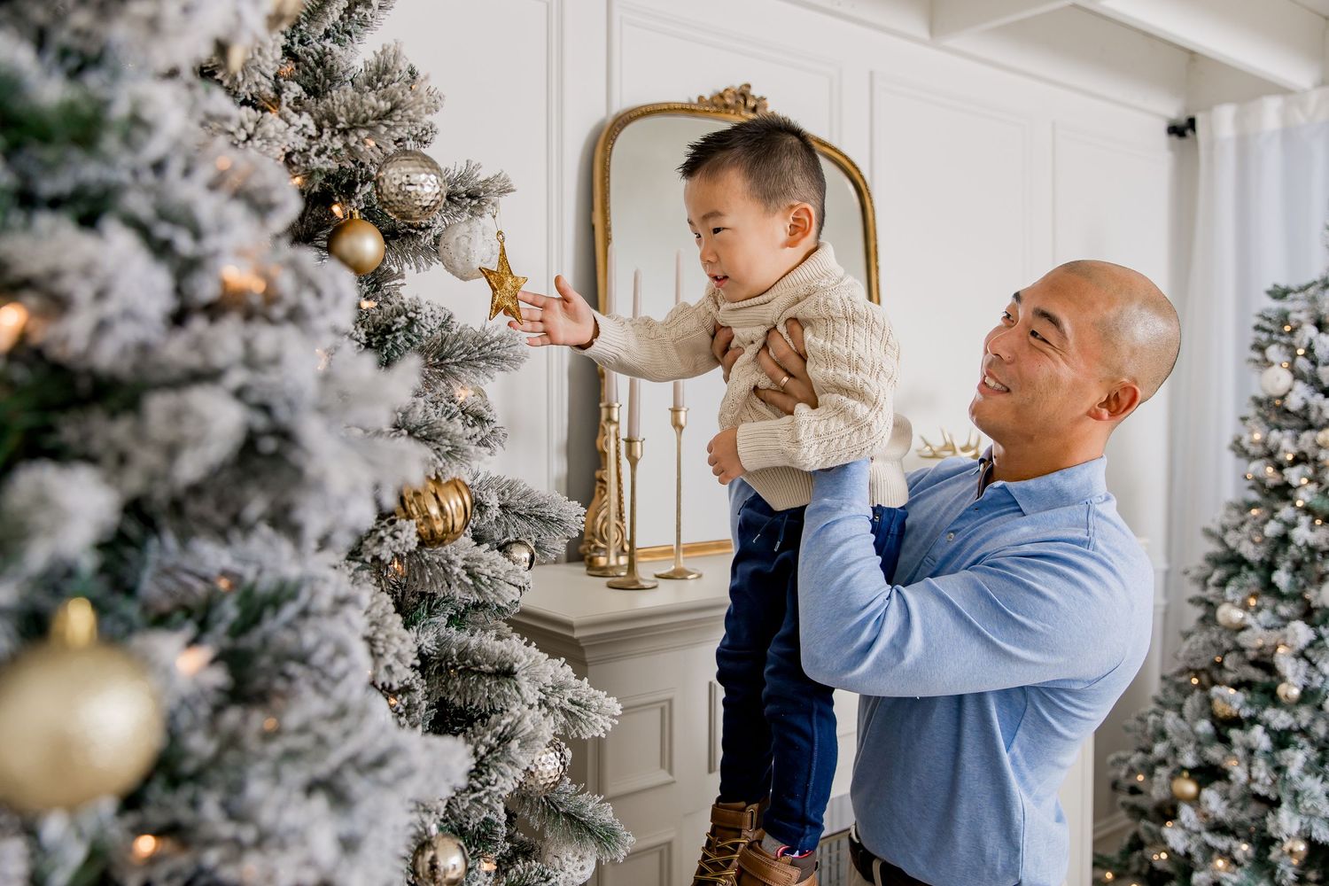 Dad lifts up toddler son to decorate a snowy white Christmas tree with ornaments in a bright festive living room setting.
