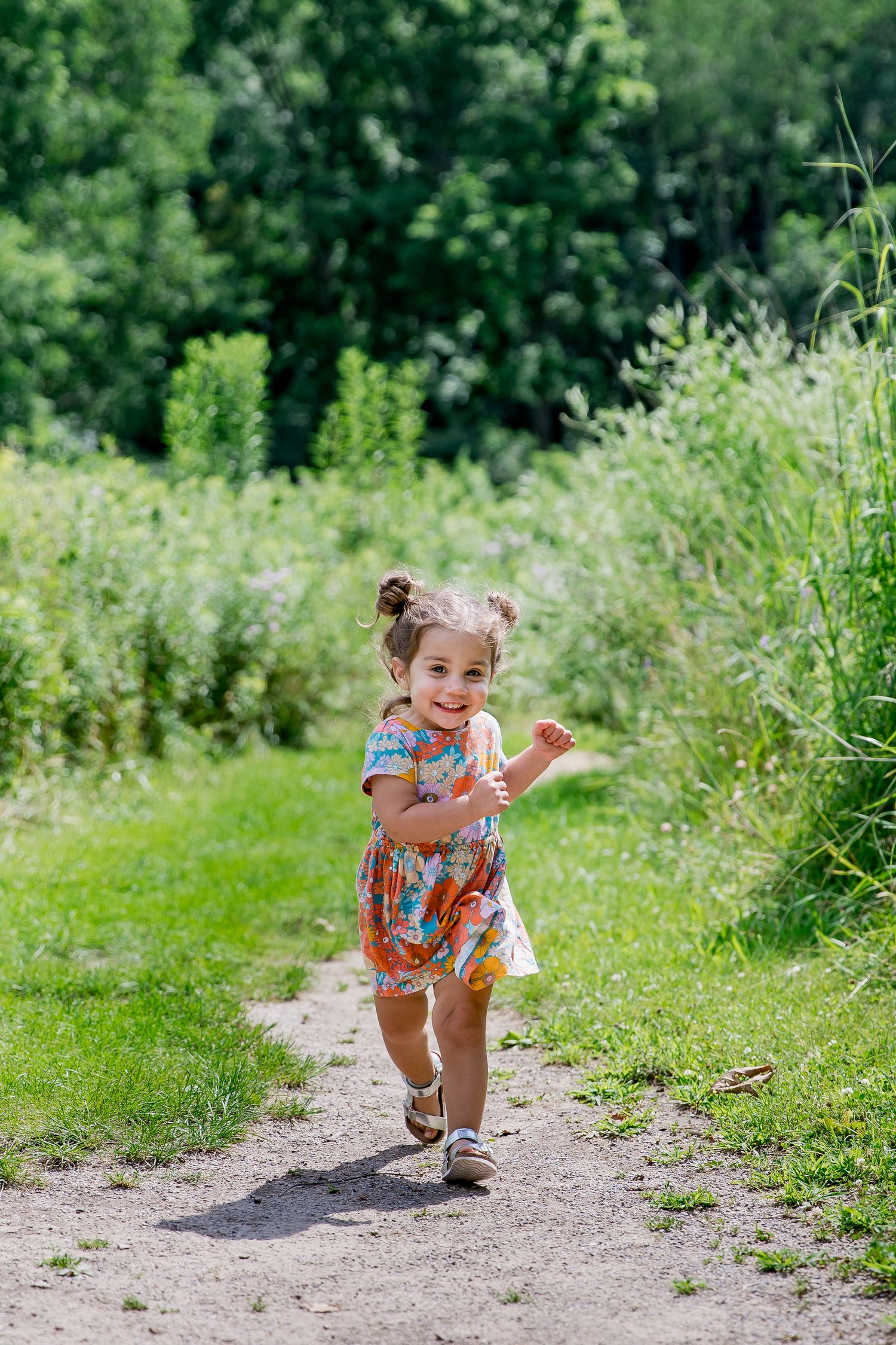 Small child in summer dress runs joyfully along a dirt path surrounded by lush green vegetation on a sunny day.