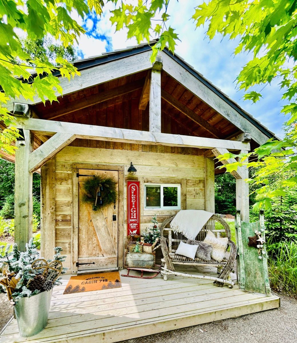 A cozy rustic cabin porch with rattan love seat, wreath on door and green maple leaves framing the view.