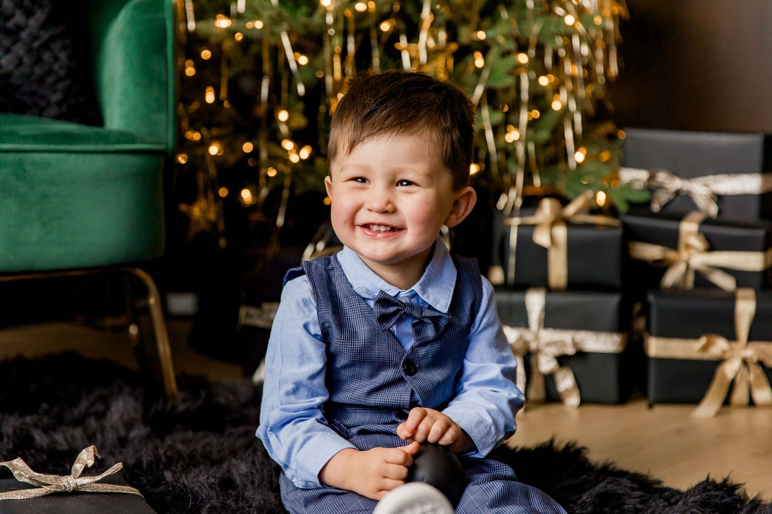A young child sits in front of a decorated Christmas tree wearing a blue vest and shirt while smiling brightly.