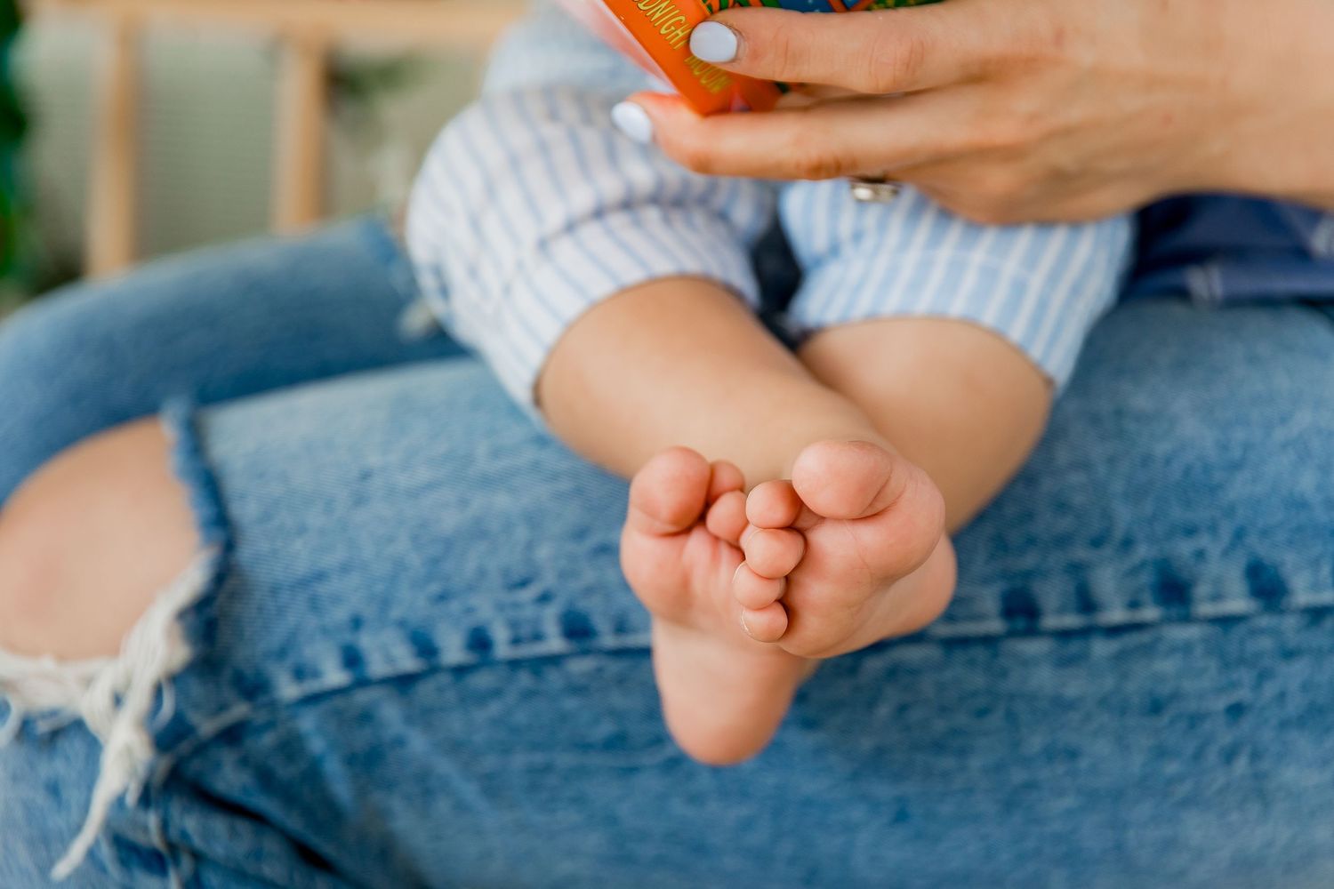 Mom in ripped blue jeans has baby sitting on her lap to read a book while baby's cute little bare feet point toward the camera in focus.