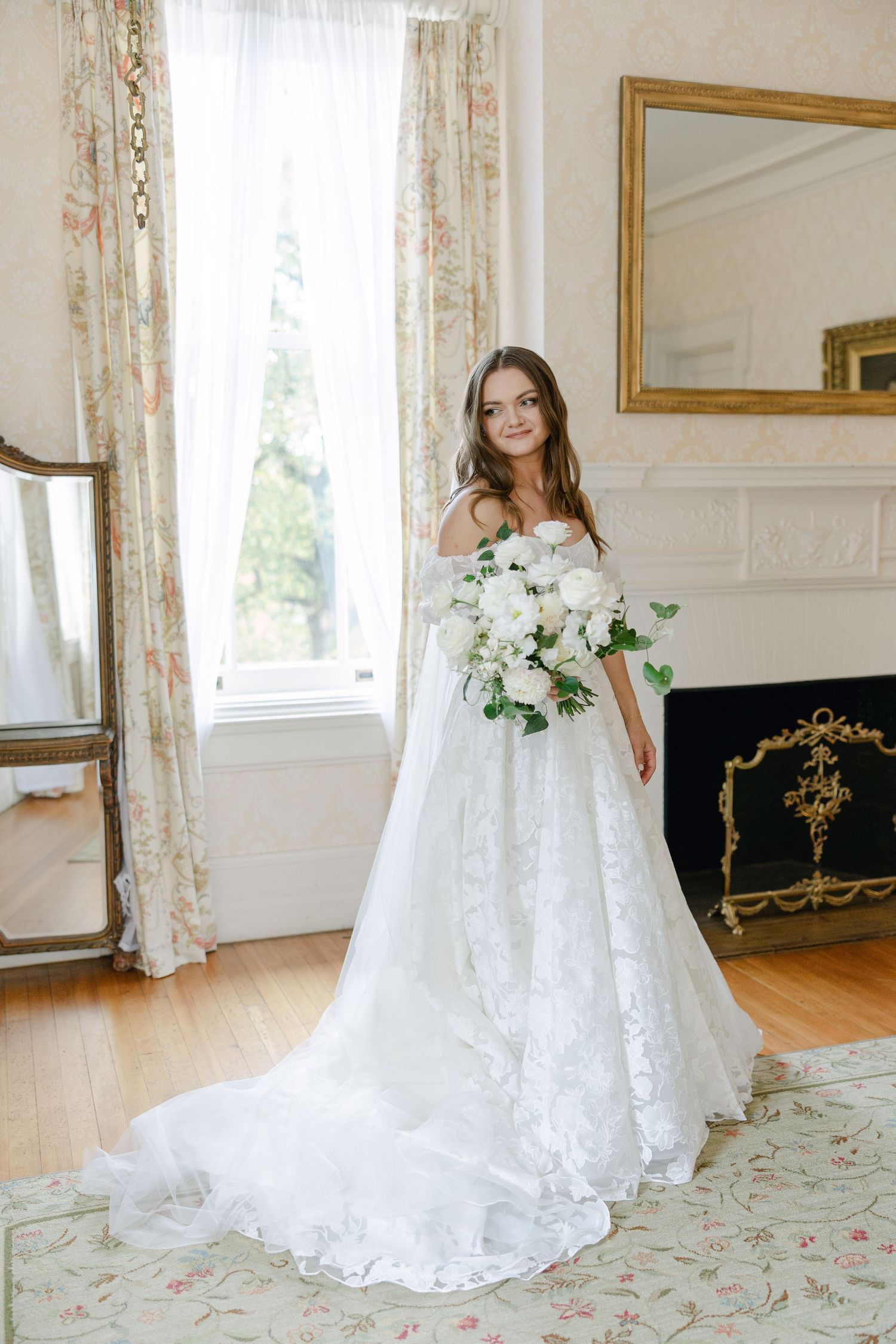 Wedding dress displayed in sunlit room with floral curtains and hardwood floors.
