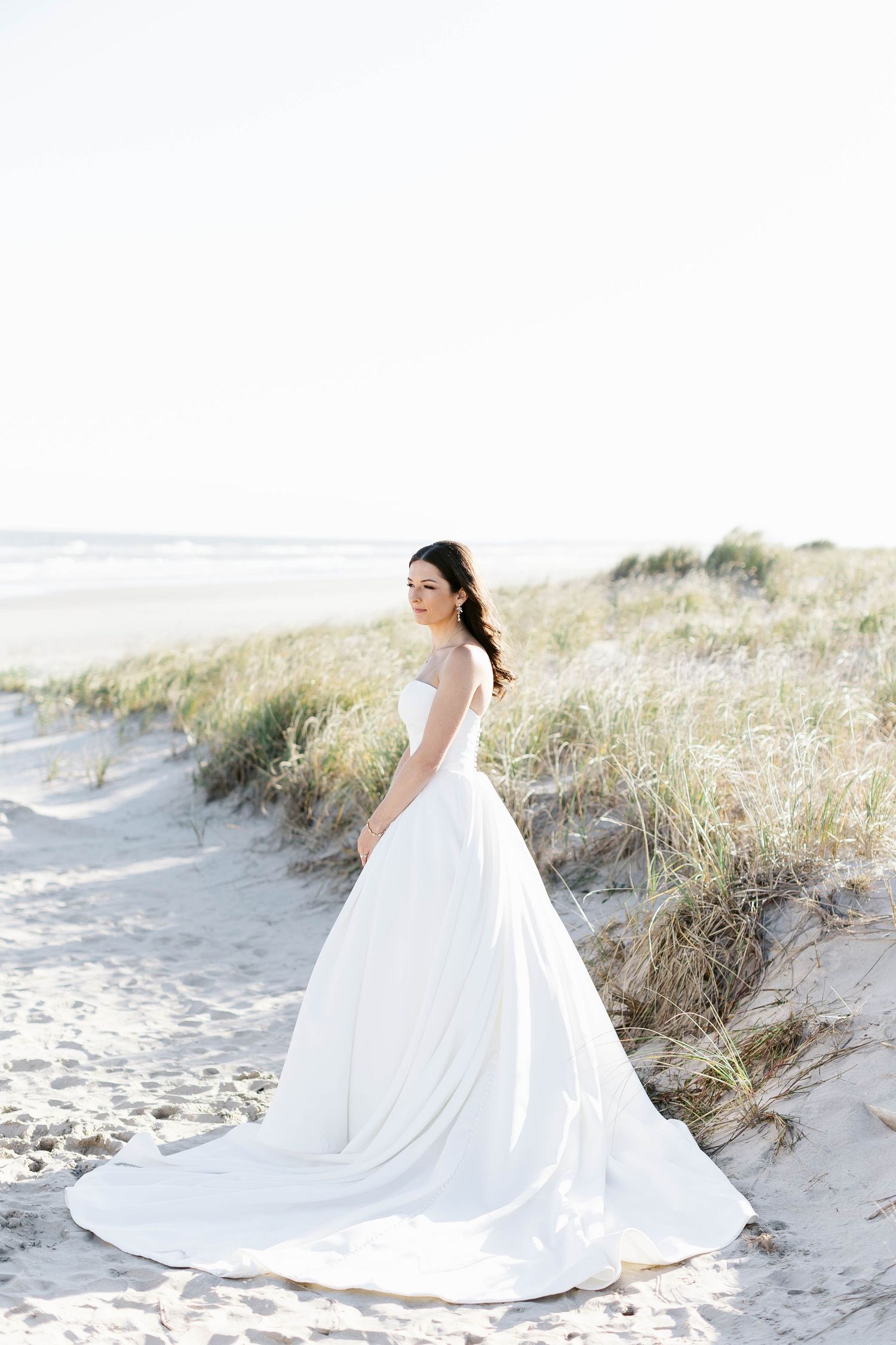 A flowing white wedding dress with long train creates dramatic silhouettes against sandy beach dunes.