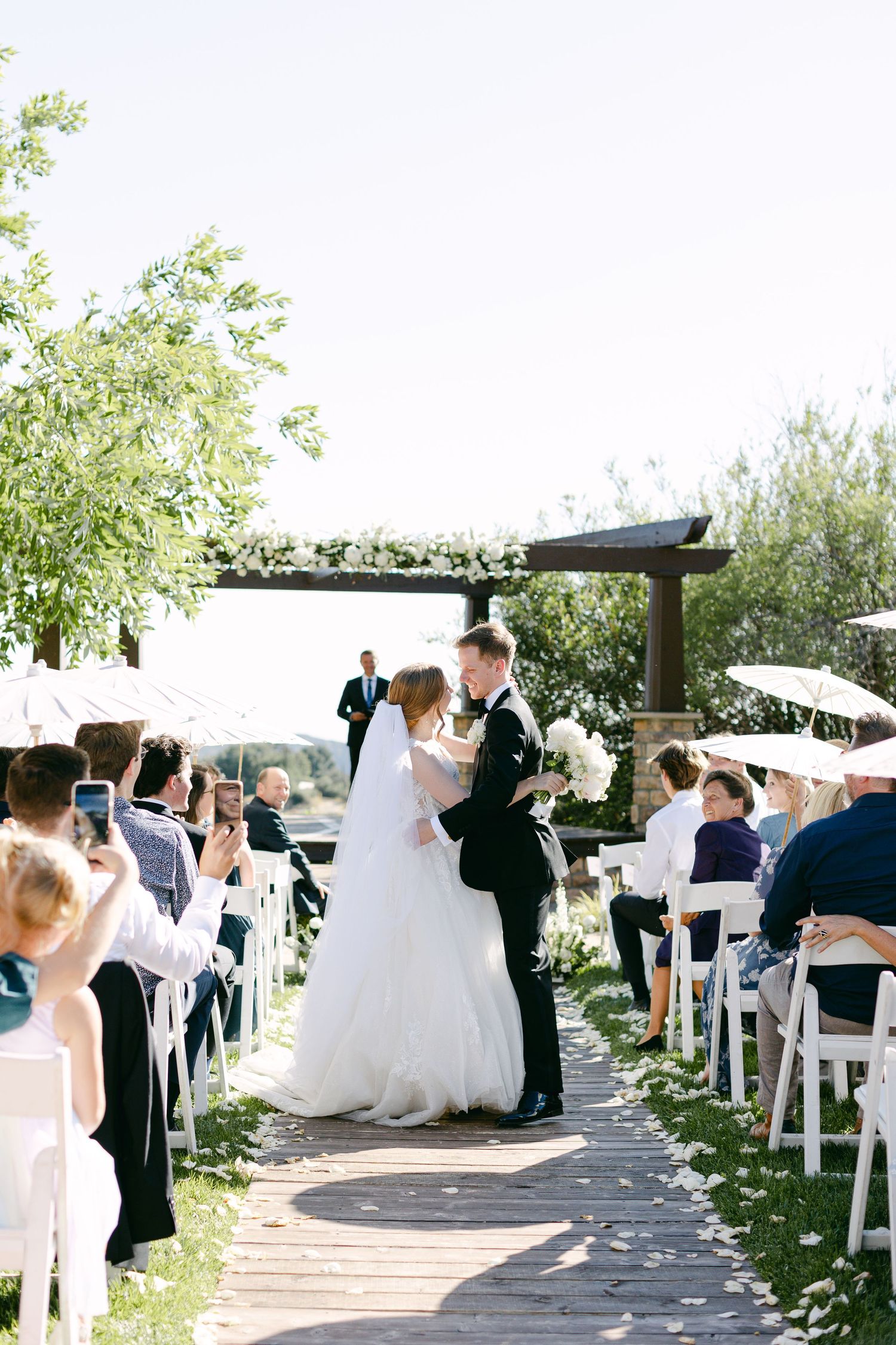 Wedding guests gather on a sunny outdoor patio decorated with white chairs and floral arrangements.