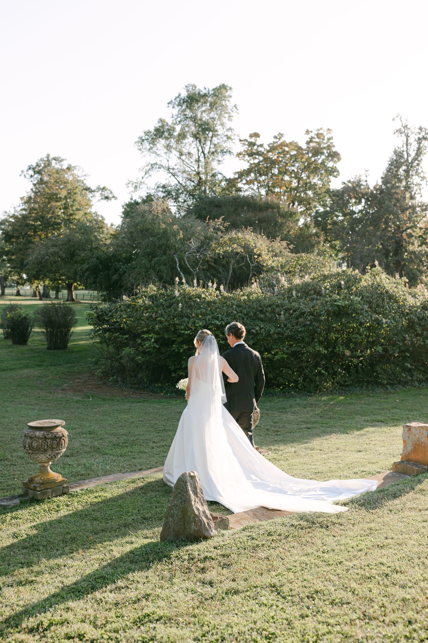 A couple strolls through a garden setting with a long white wedding dress train trailing behind them.