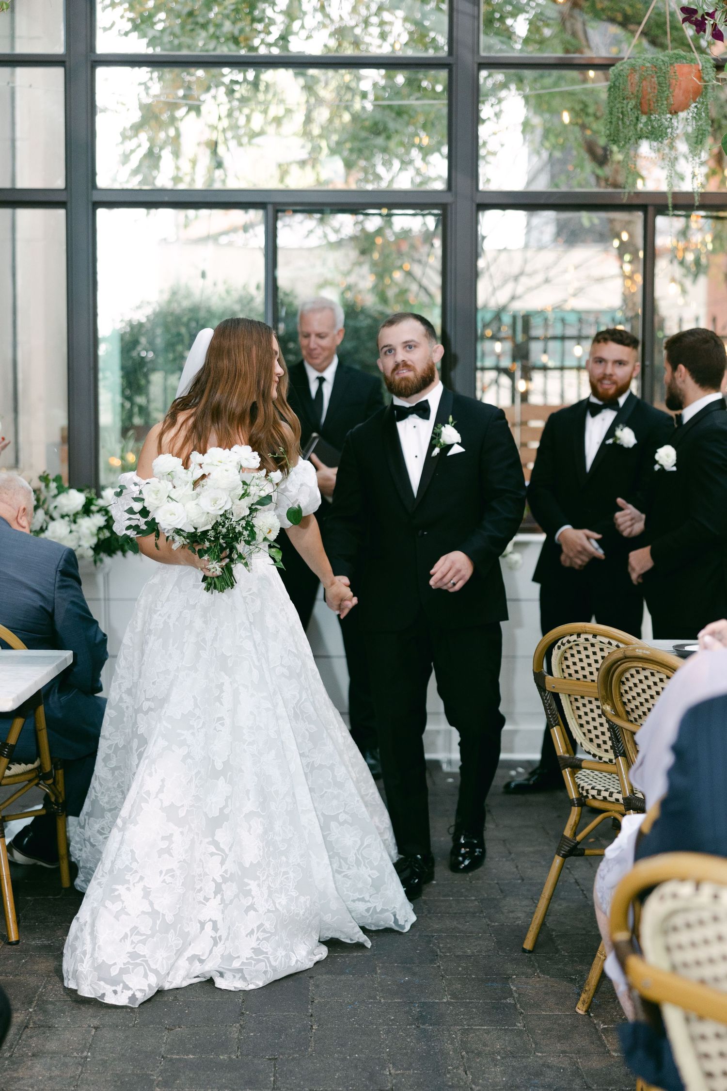 A wedding ceremony in a glass-walled venue with guests seated in bistro chairs.