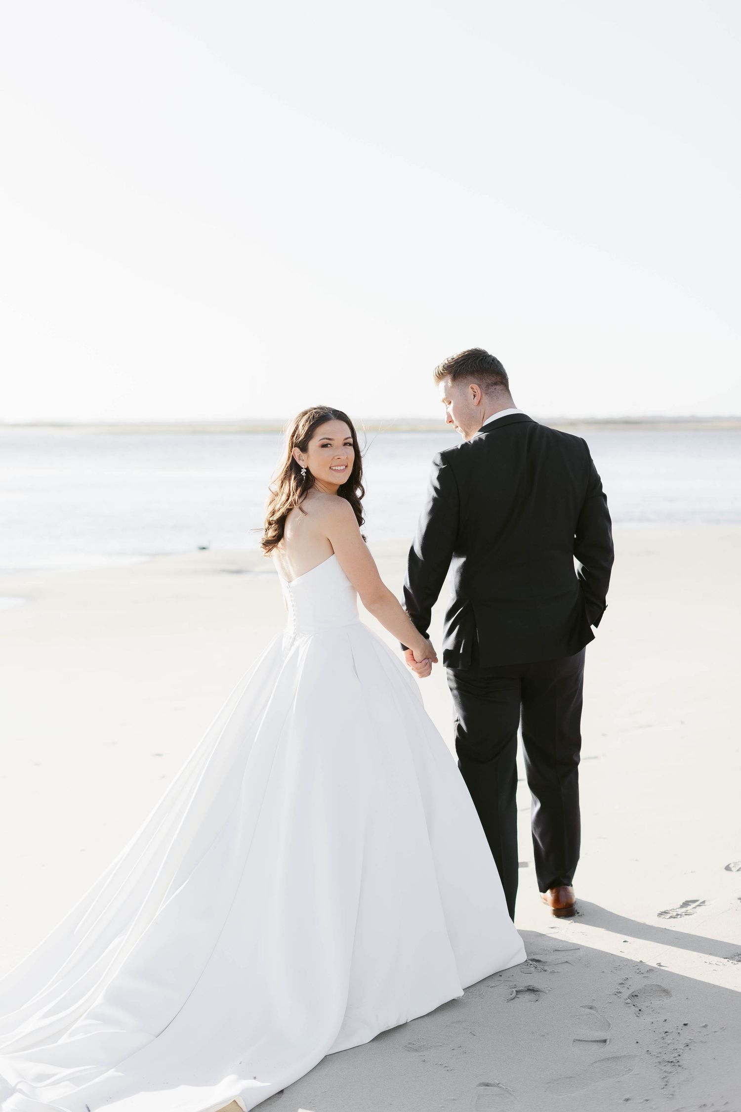 Wedding couple walks hand in hand along pristine beach with dramatic white dress trailing behind.