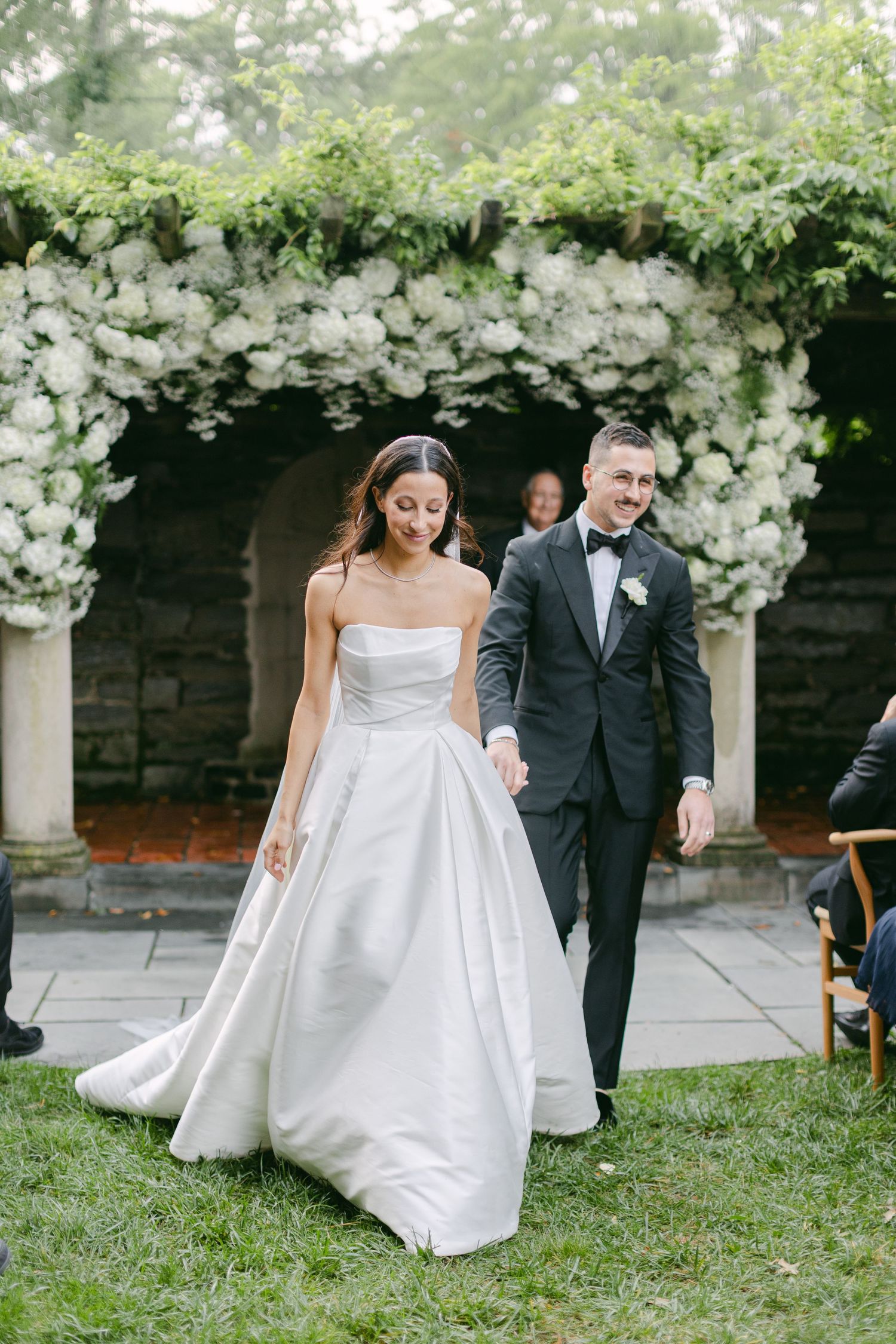 Wedding couple walking under white floral arch decorations in an outdoor garden setting.