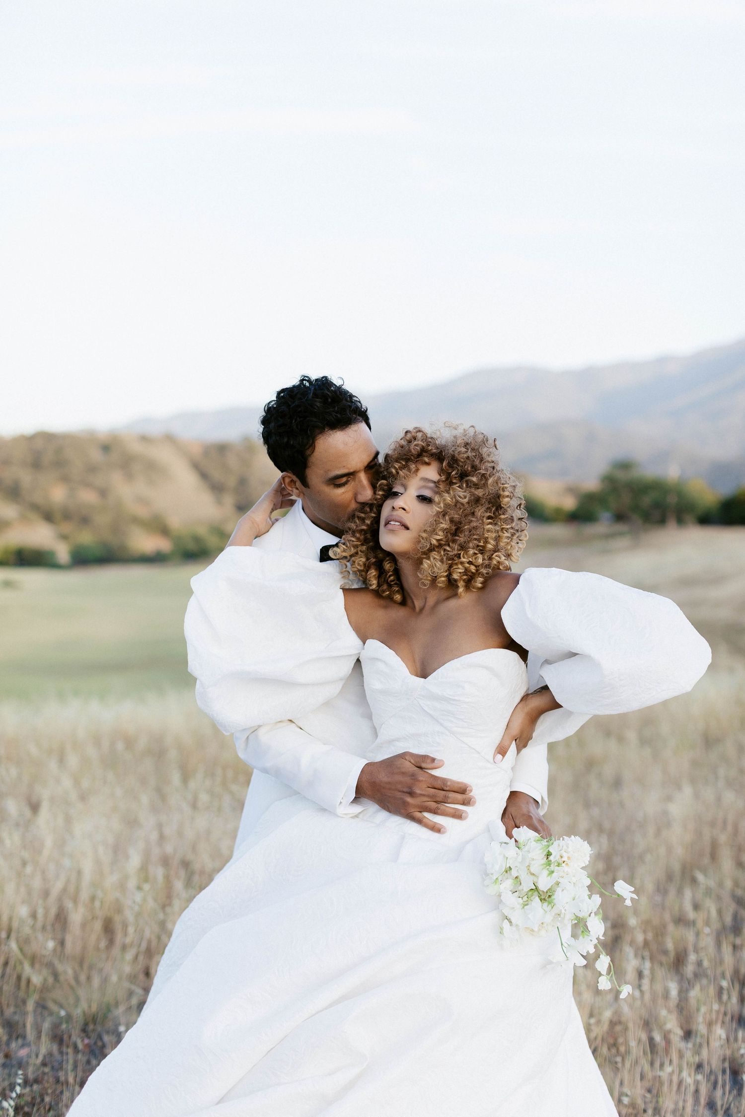 A romantic wedding portrait of a couple in white attire embracing in a golden field with mountains in the background.