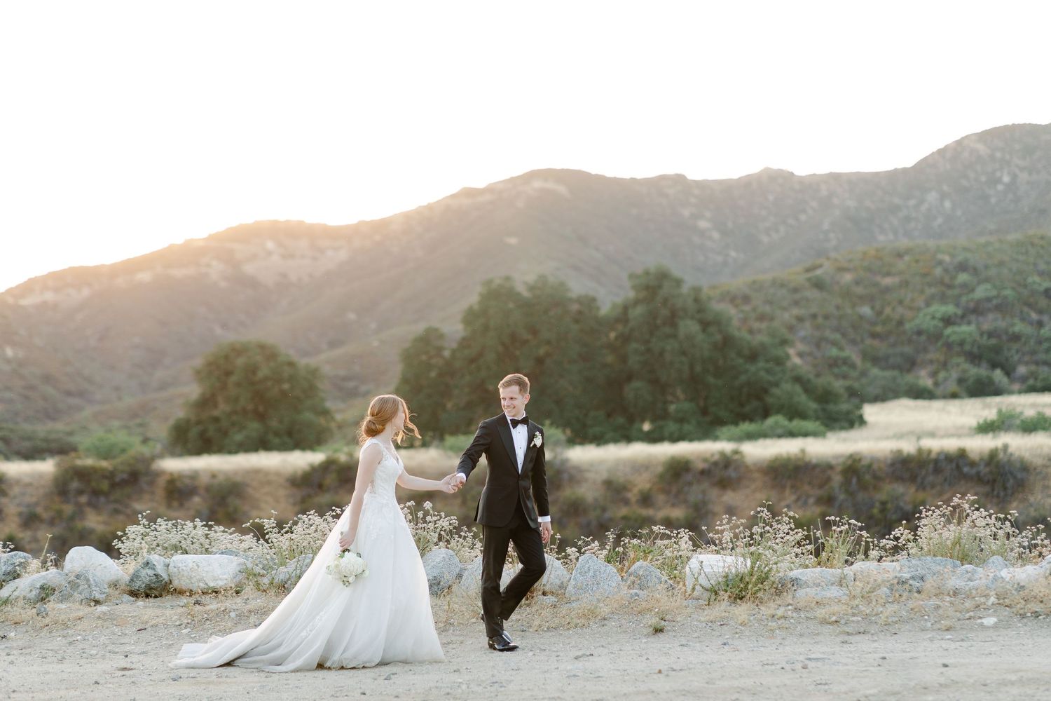 Couple in wedding attire walking through golden field at sunset with mountains in background.