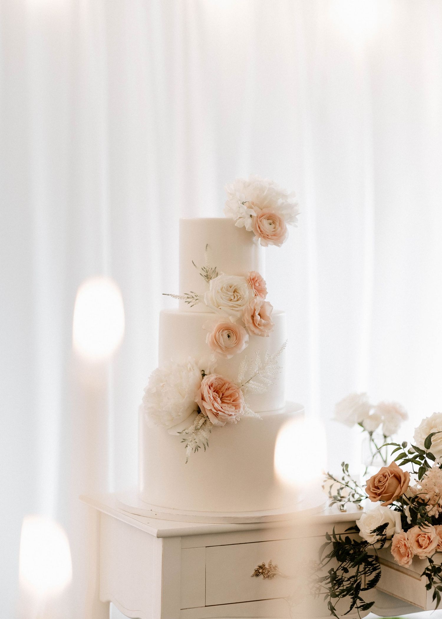 White wedding cake decorated with delicate peach roses and greenery against a light backdrop.