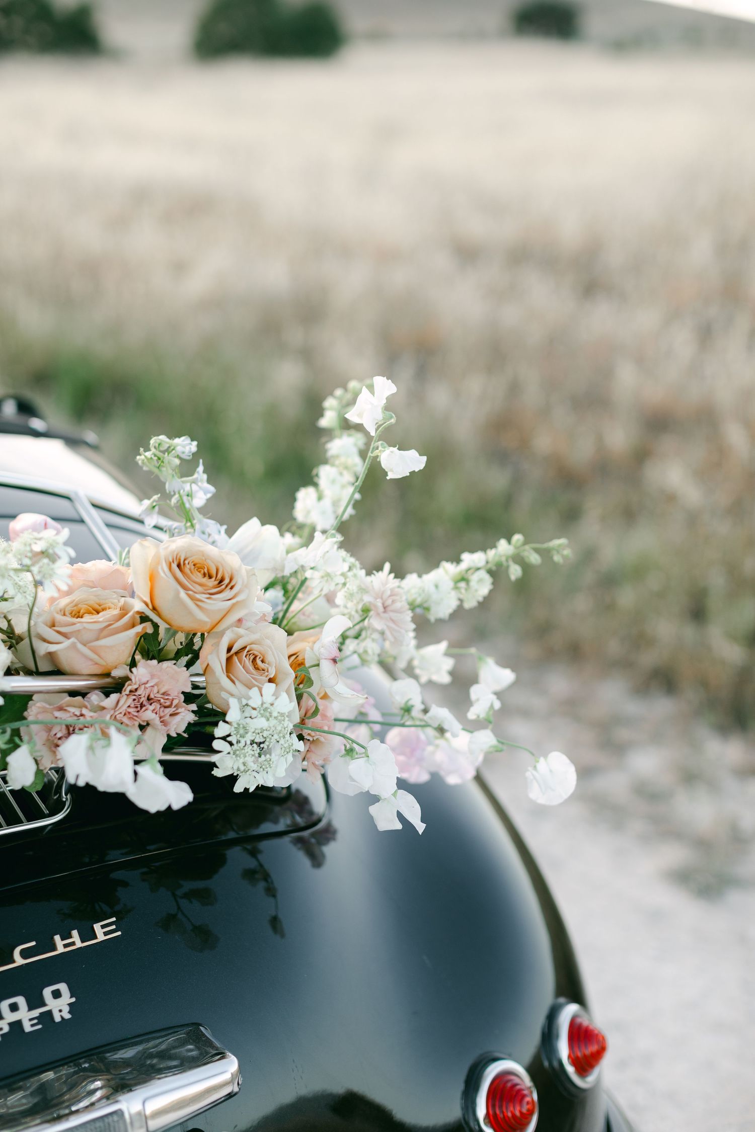 Delicate peach roses and white flowers adorn the trunk of a vintage black Porsche.