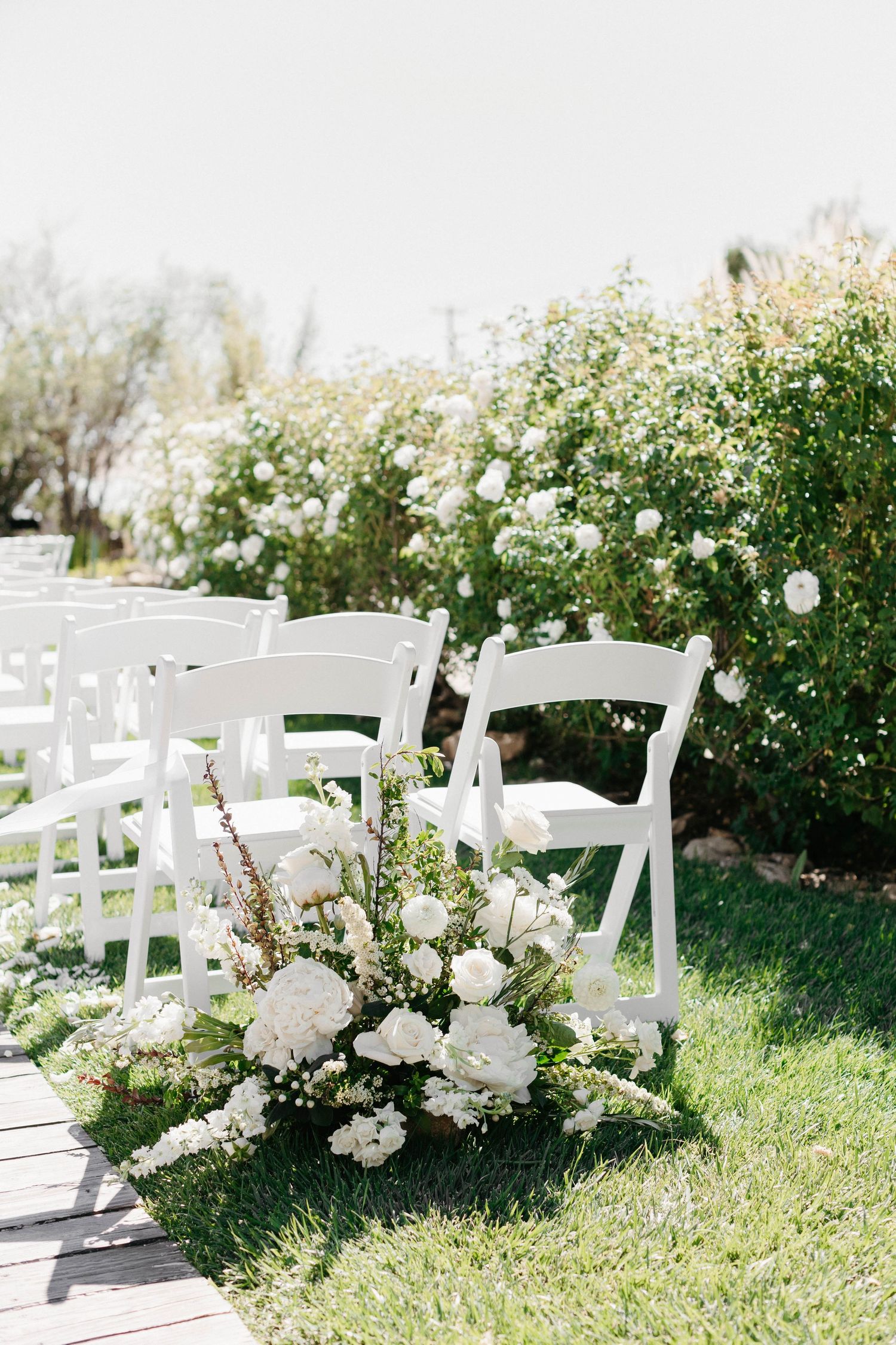 White garden chairs and floral arrangements line the outdoor wedding ceremony aisle.