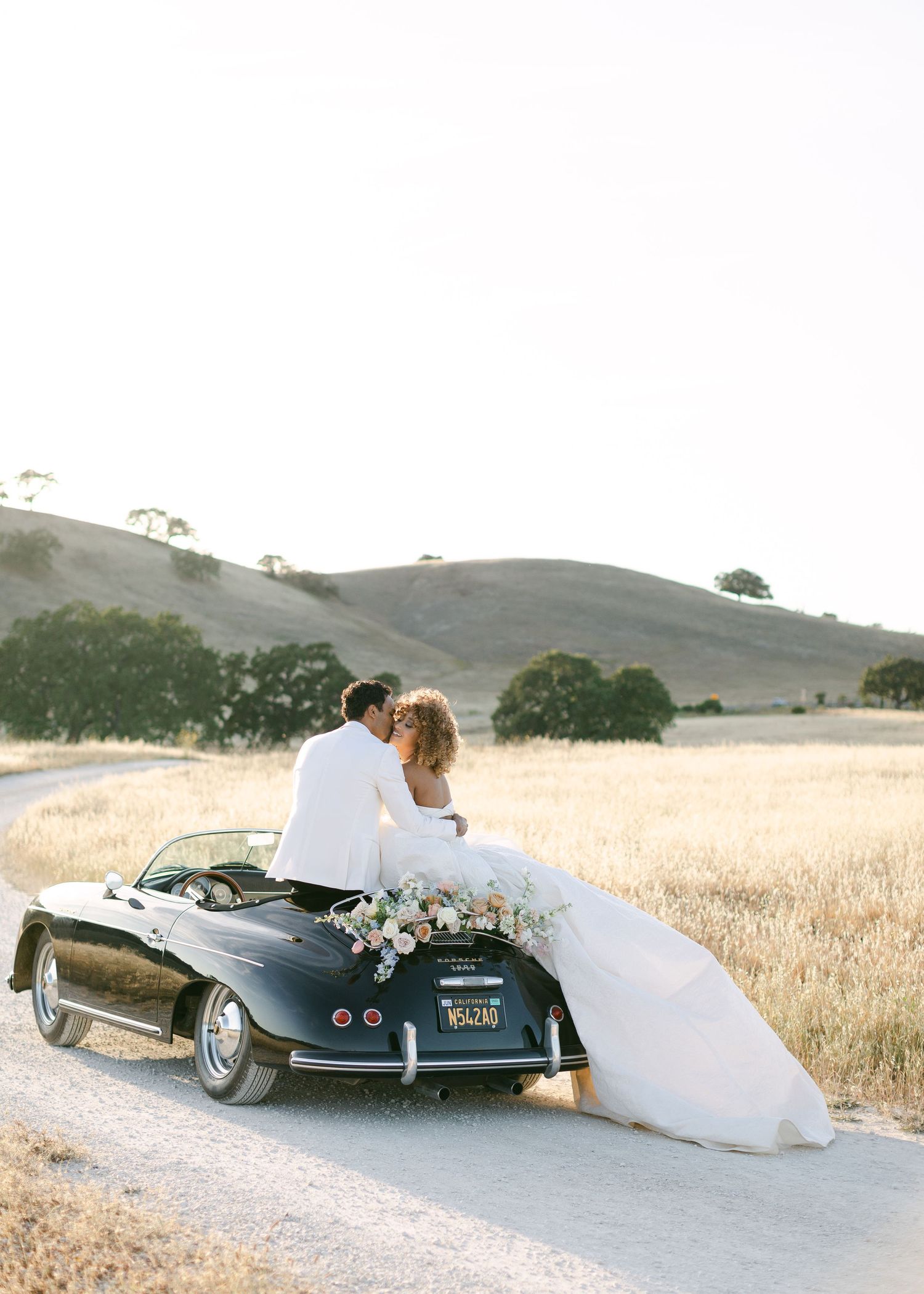 A classic black vintage car decorated with flowers serves as a wedding getaway vehicle in a rustic field.