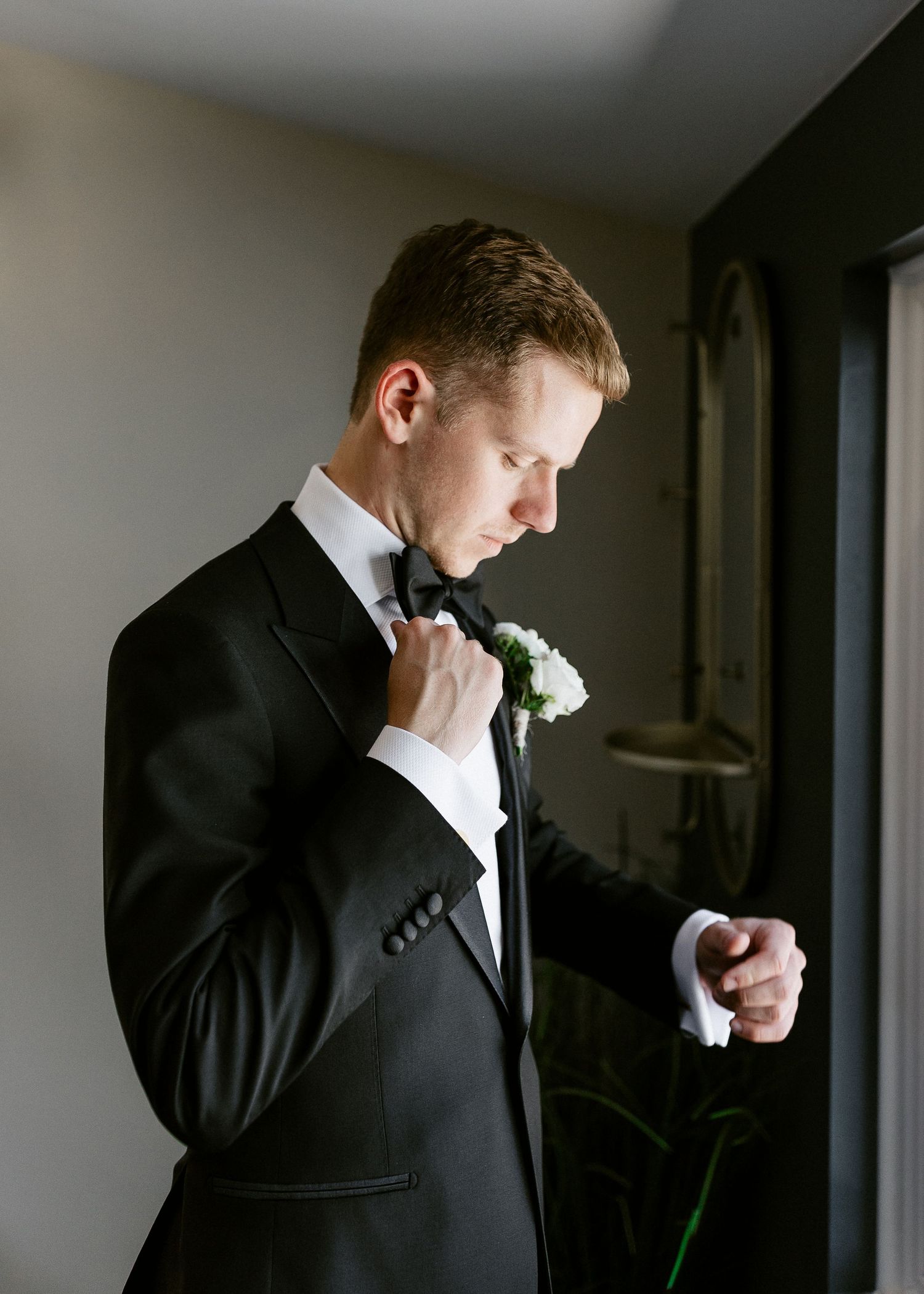 A groom in a black tuxedo adjusts his boutonniere near a window.