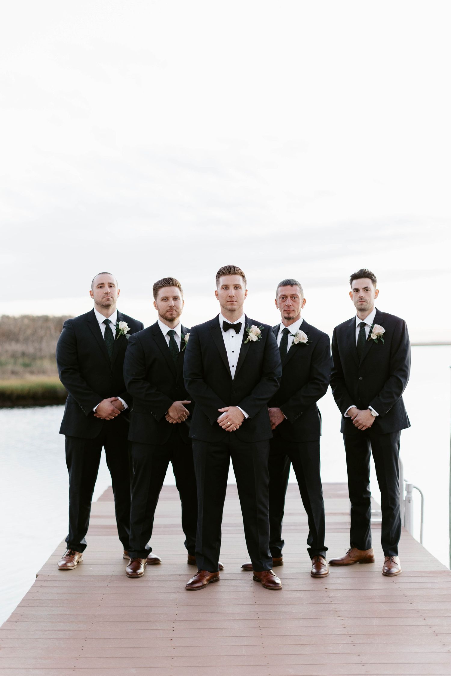 Groomsmen in black suits pose playfully on wooden dock by waterfront.